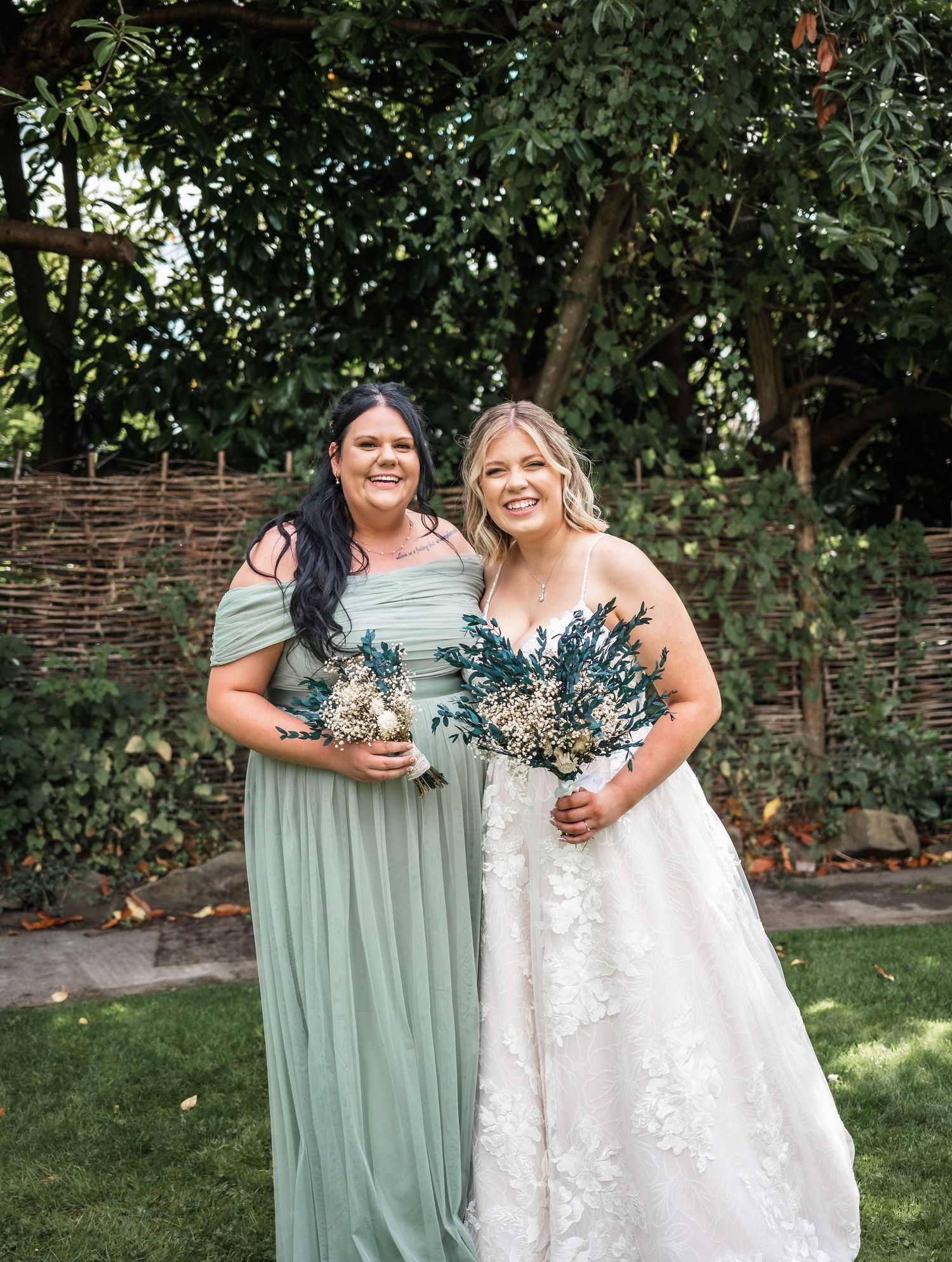 Bride in white lace gown and bridesmaid in sage green dress pose with bouquets in garden setting.