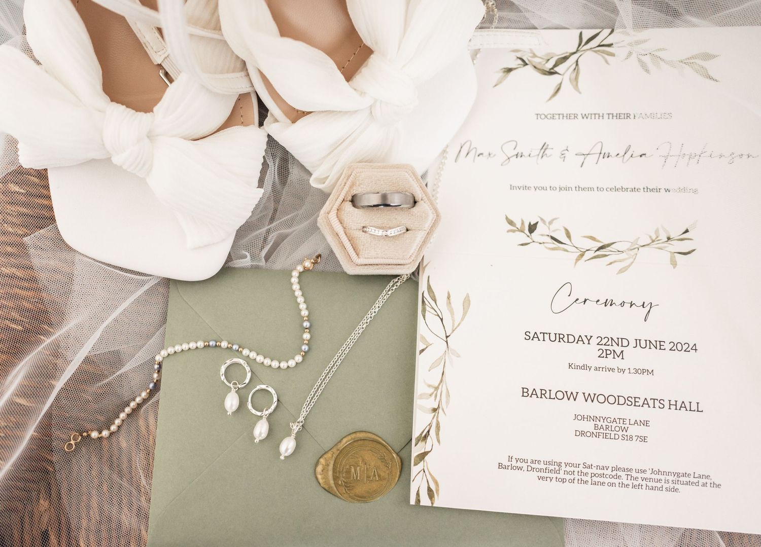 Wedding invitation and jewelry details with white floral elements and sage green accents on a wooden surface.