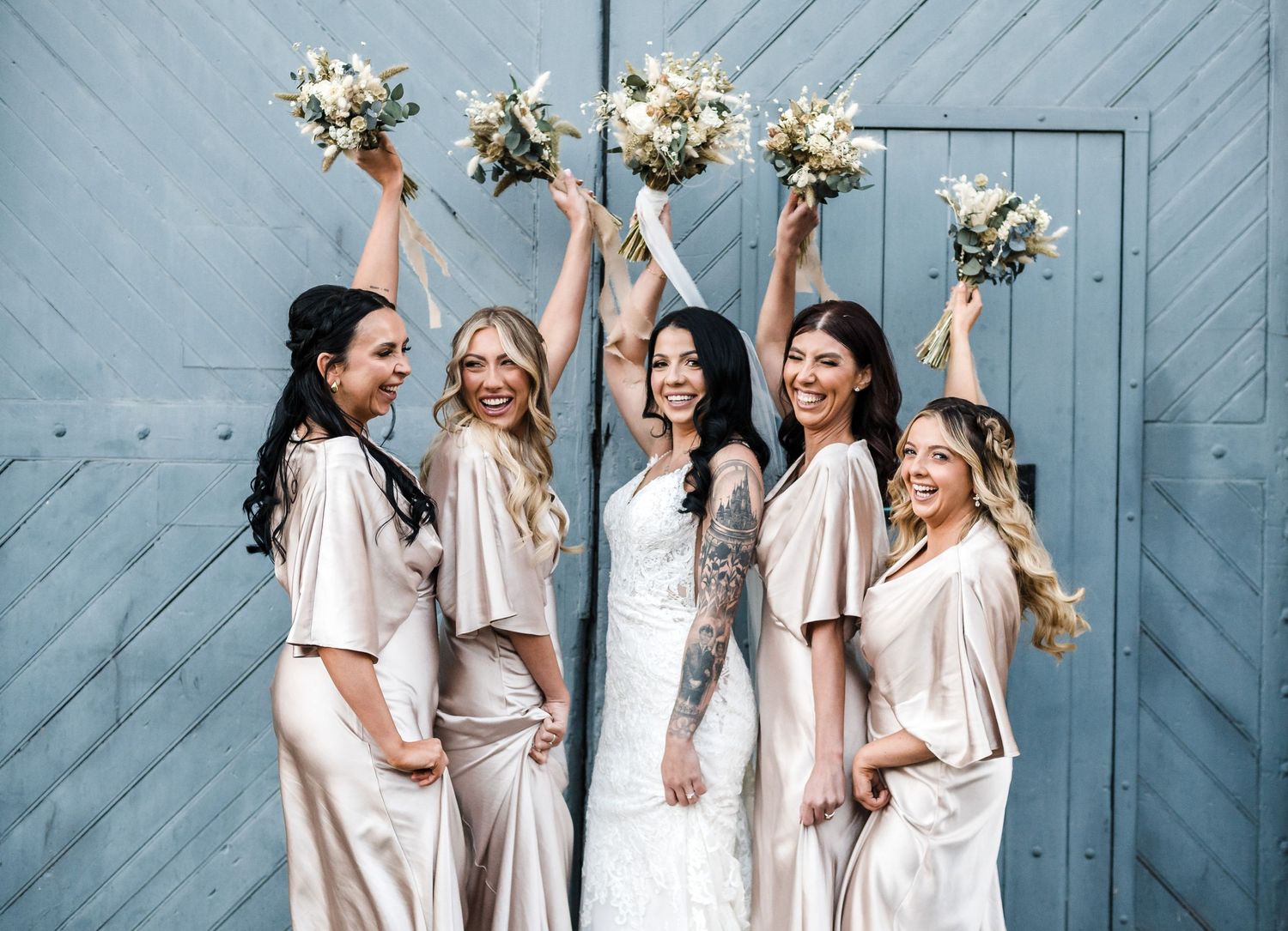 Bridesmaids in silk dresses raise bouquets in celebration against blue door.