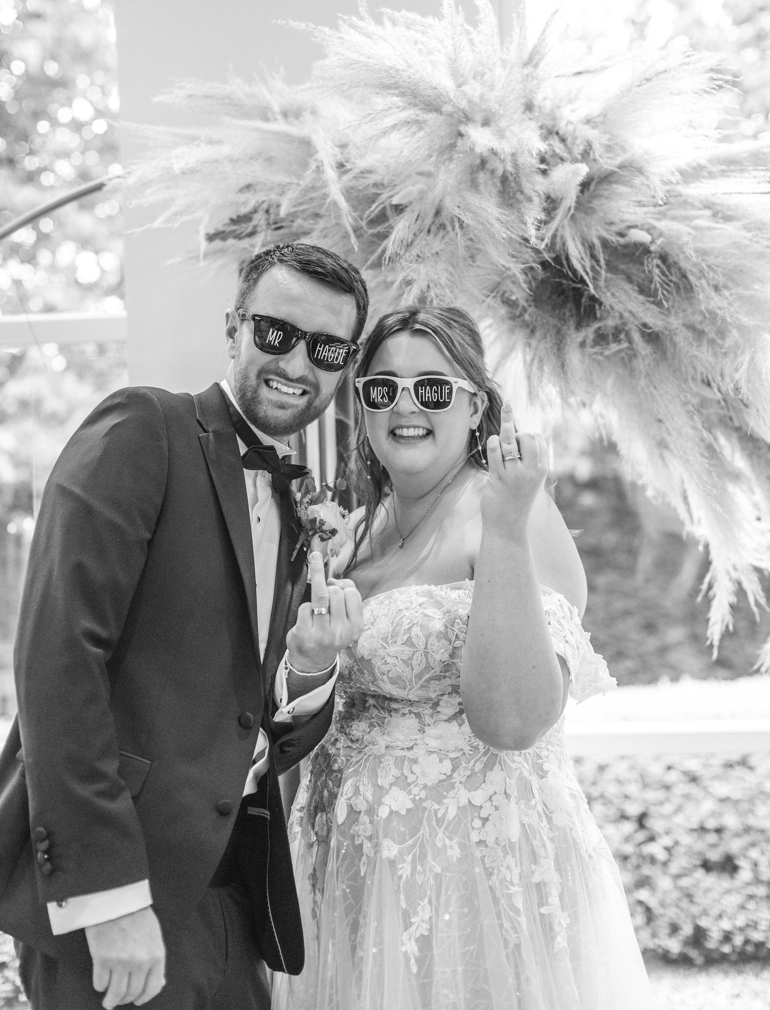A wedding couple in sunglasses and formal attire smiling together with a pampas grass bouquet in black and white.