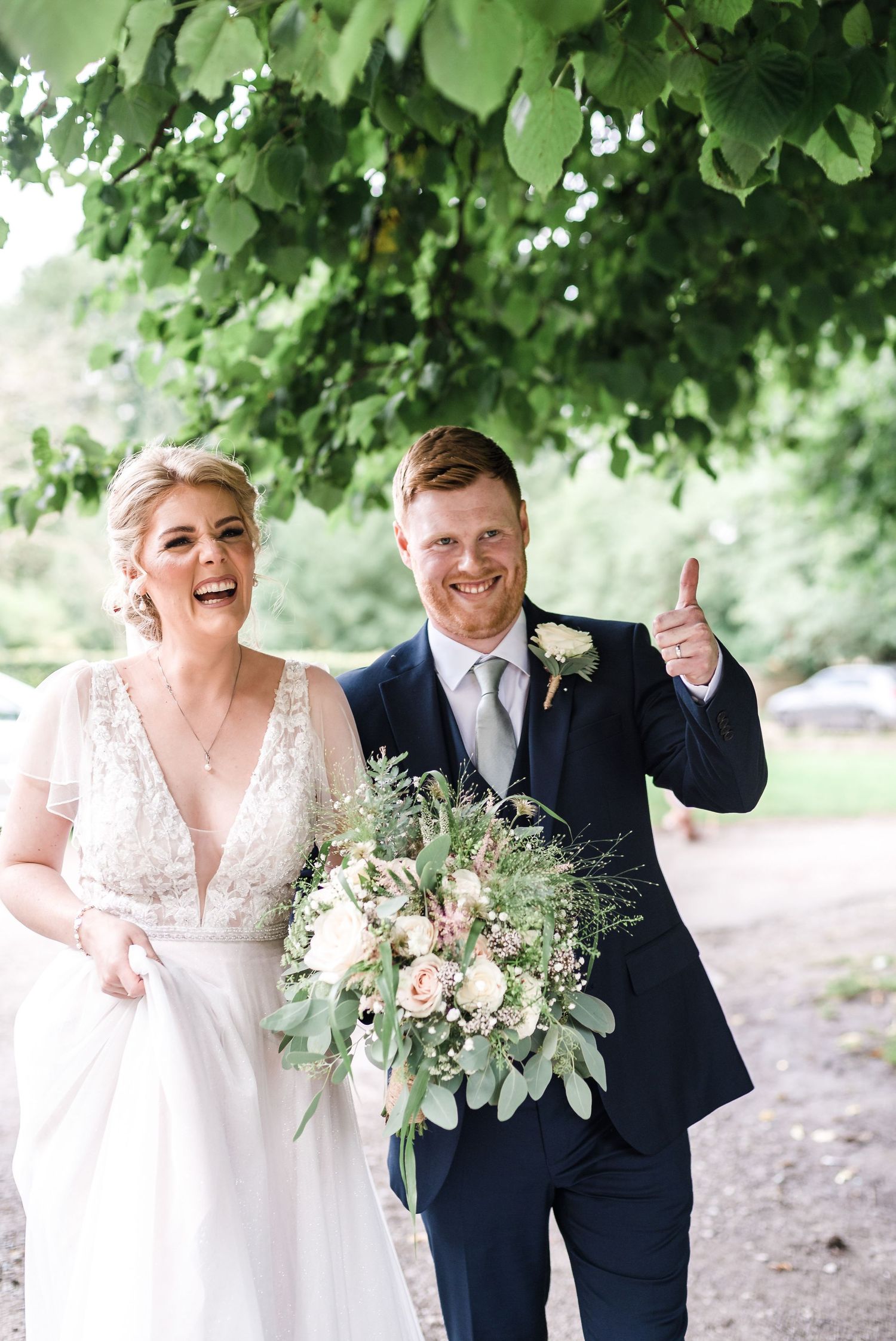 Joyful wedding couple sharing a laugh on a garden path with fresh floral bouquet.