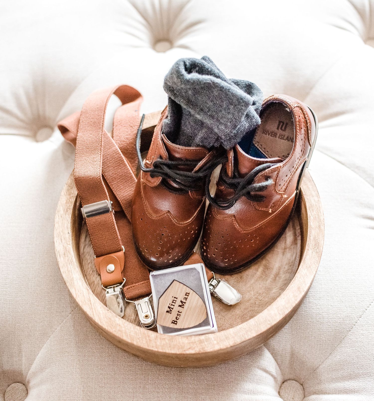 Brown leather dress shoes and gray socks arranged in wooden bowl on tufted ottoman.