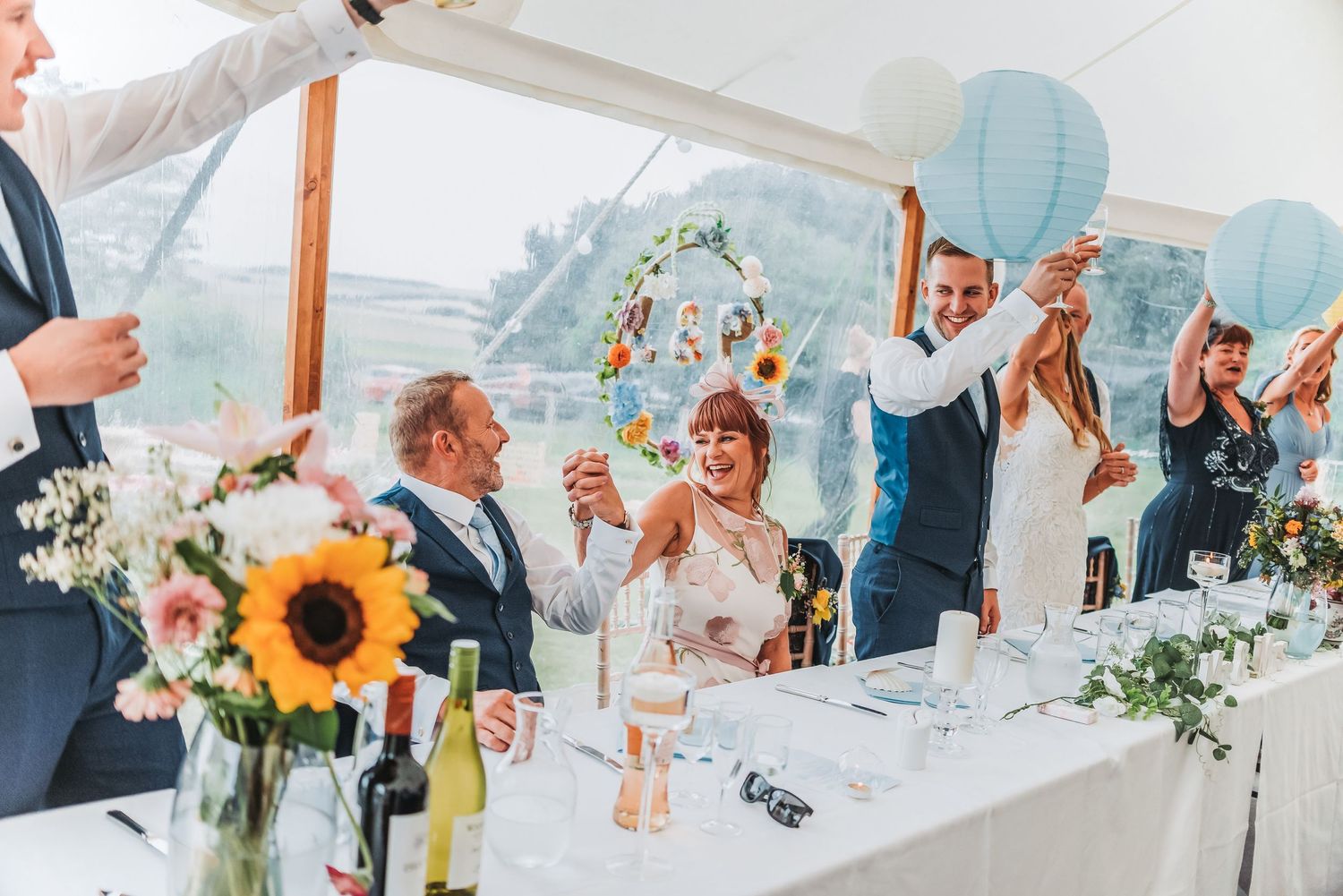 Wedding reception celebration under white tent with sunflower decorations and blue paper lanterns.