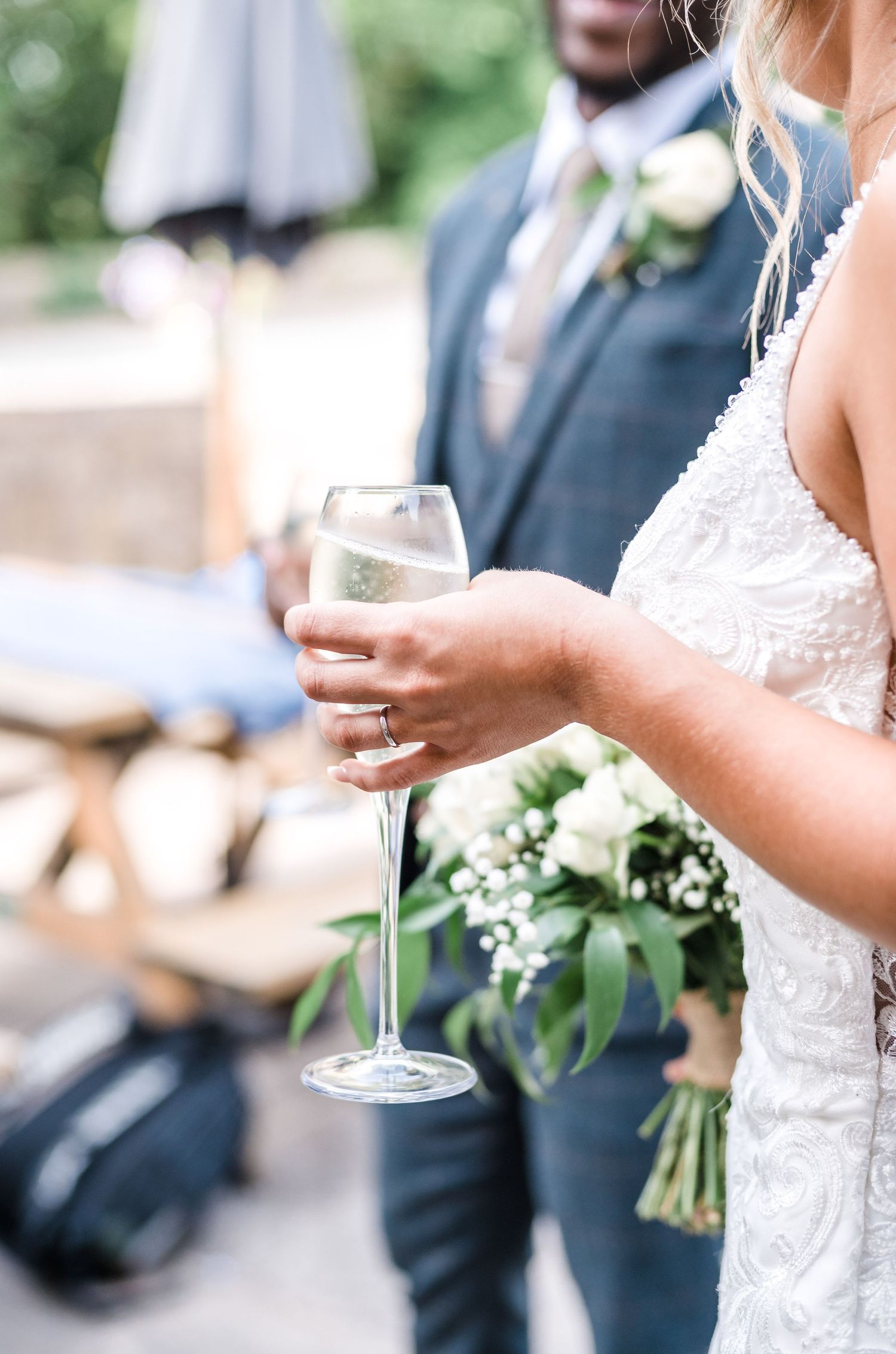 Bride holding champagne glass with white bouquet against blurred background.