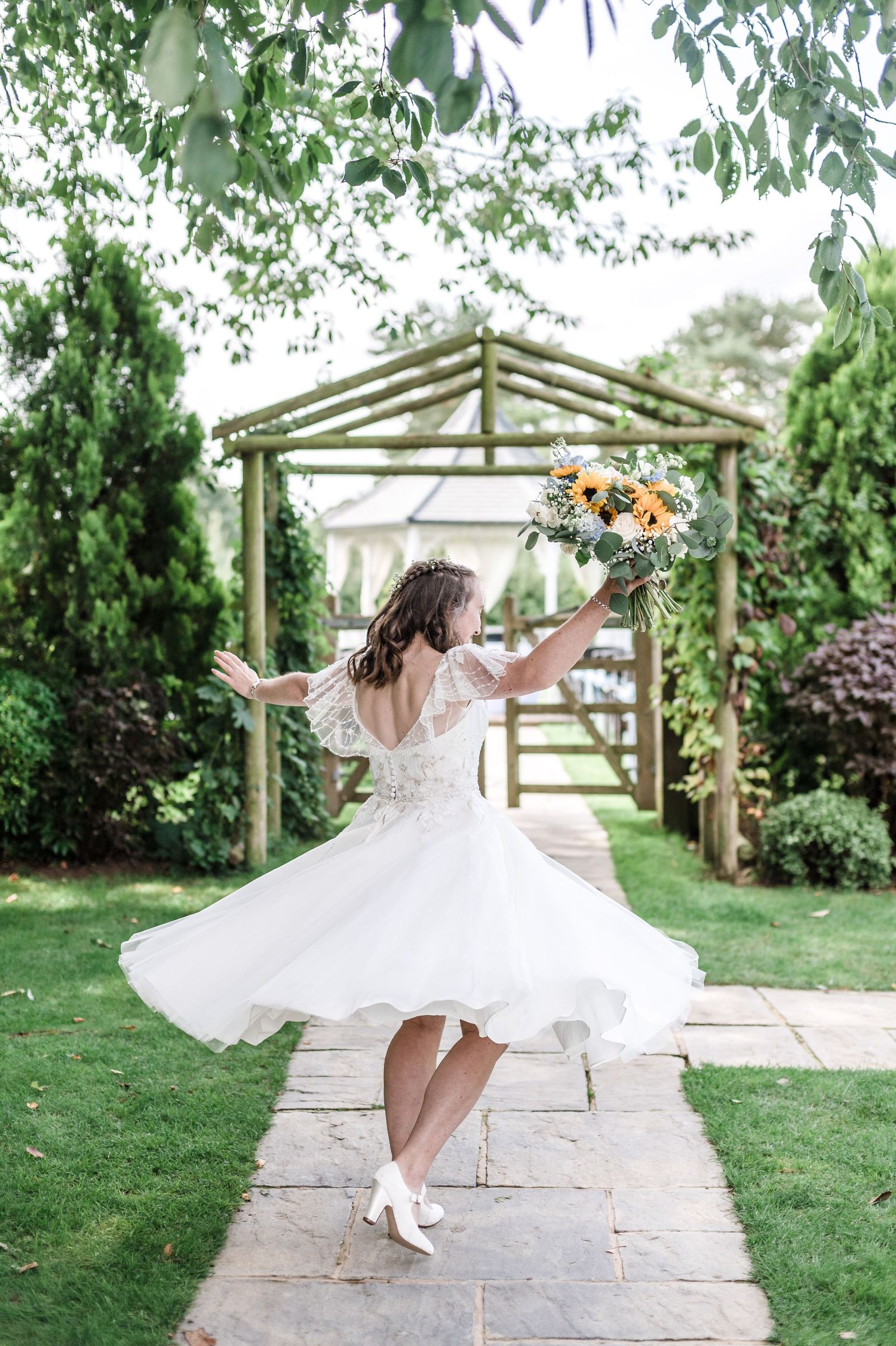 Bride in short wedding dress twirls while holding bouquet under wooden garden arbor.