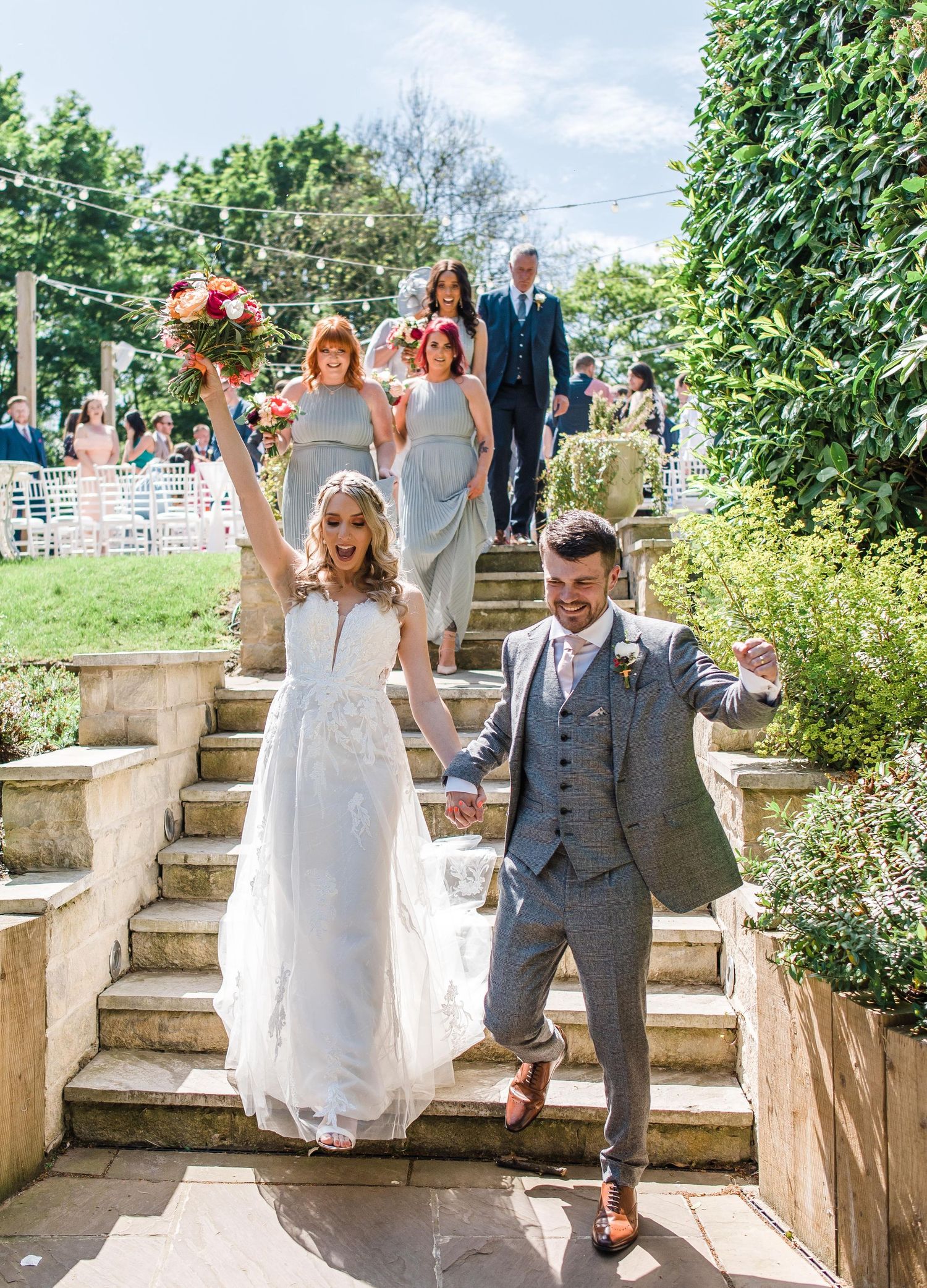 Wedding party descending stone garden steps surrounded by greenery on a sunny day.