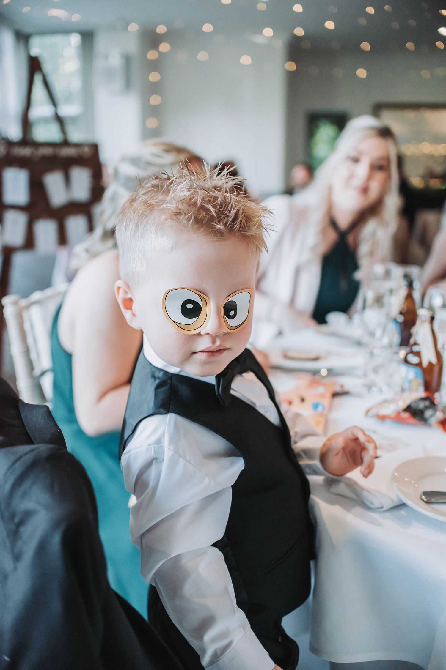Toddler in formal attire with playful expression at wedding reception table.