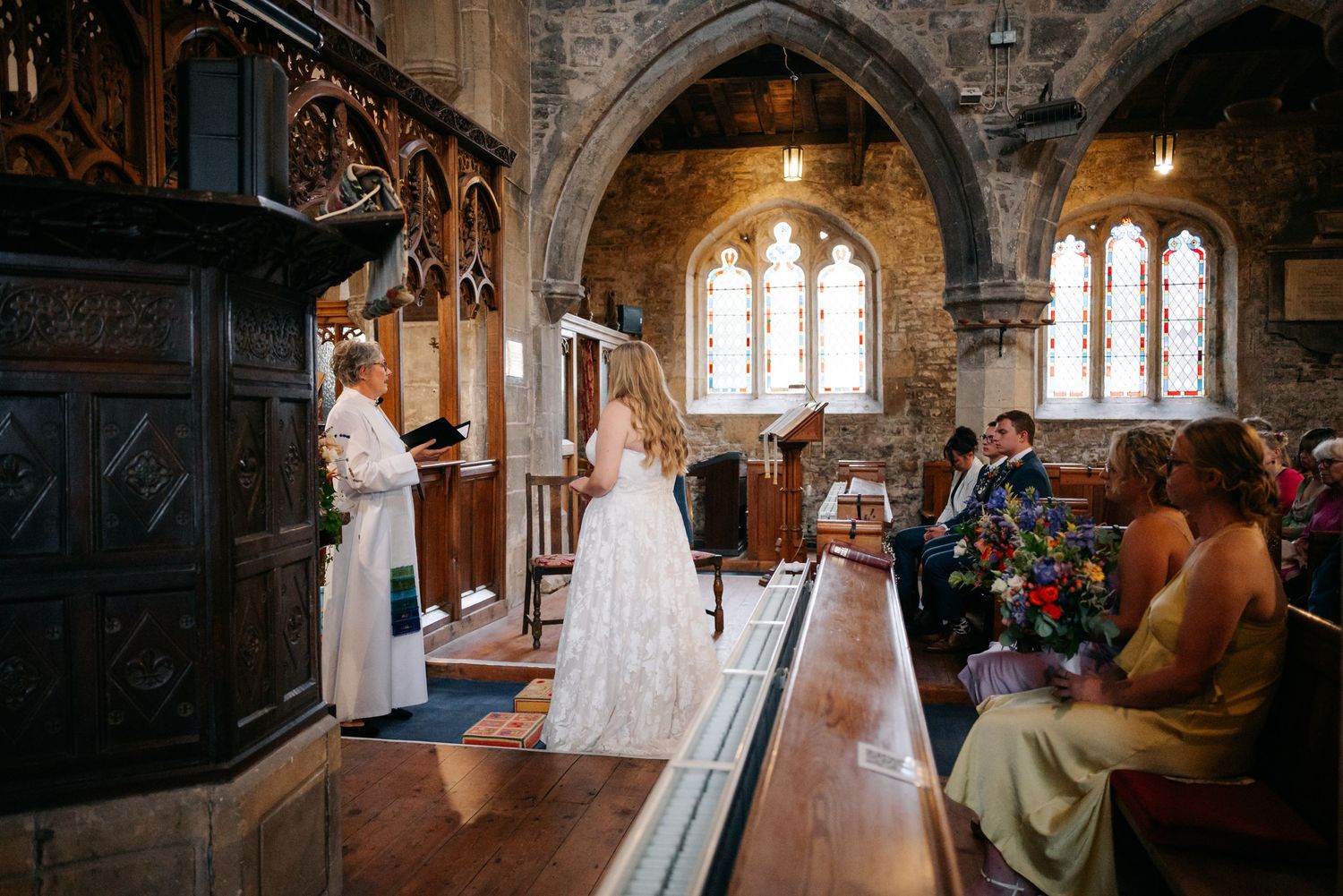 A wedding ceremony takes place in a historic stone church with sunlight streaming through stained glass windows.