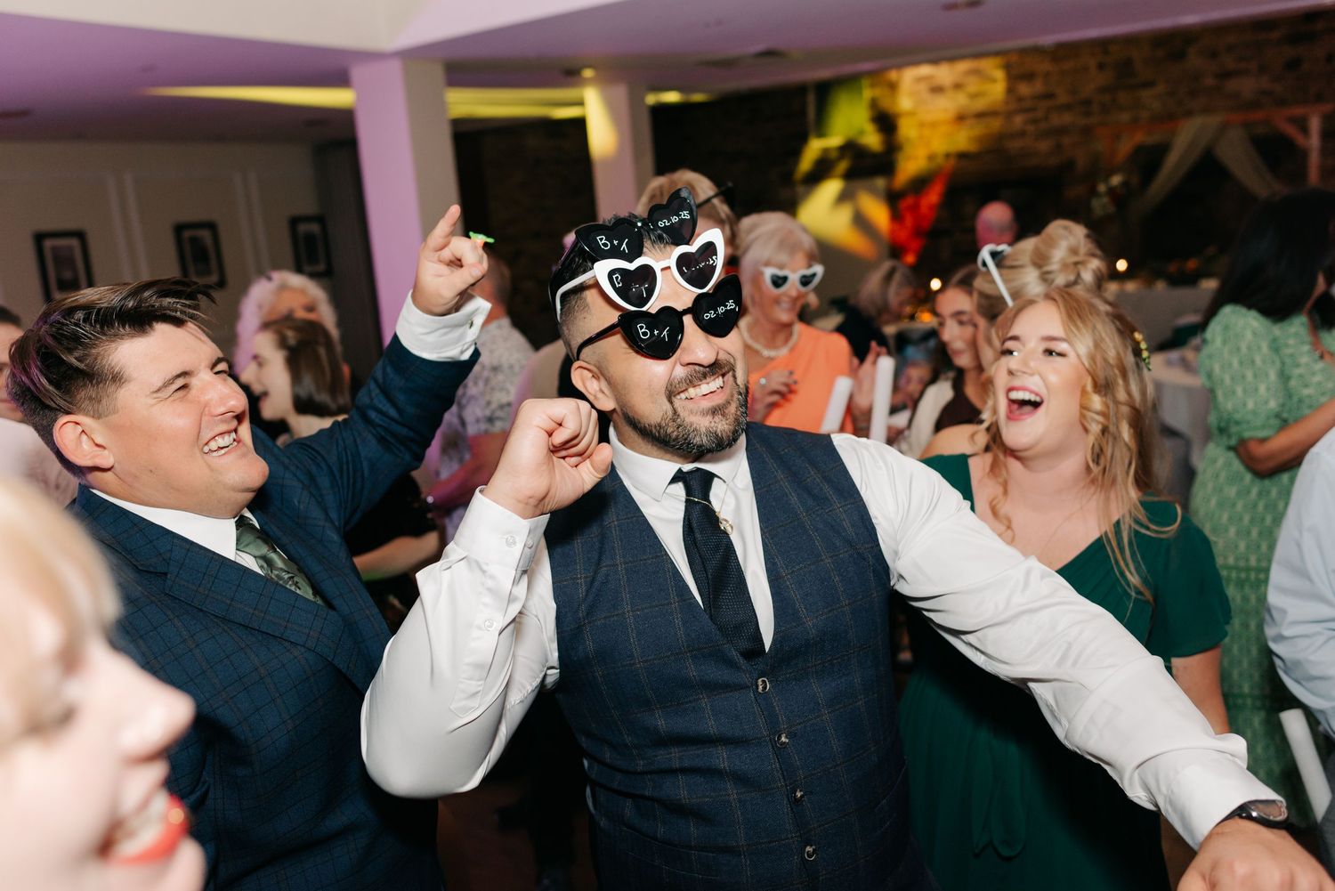 Wedding party dancing and celebrating with sunglasses in a dimly lit reception venue.