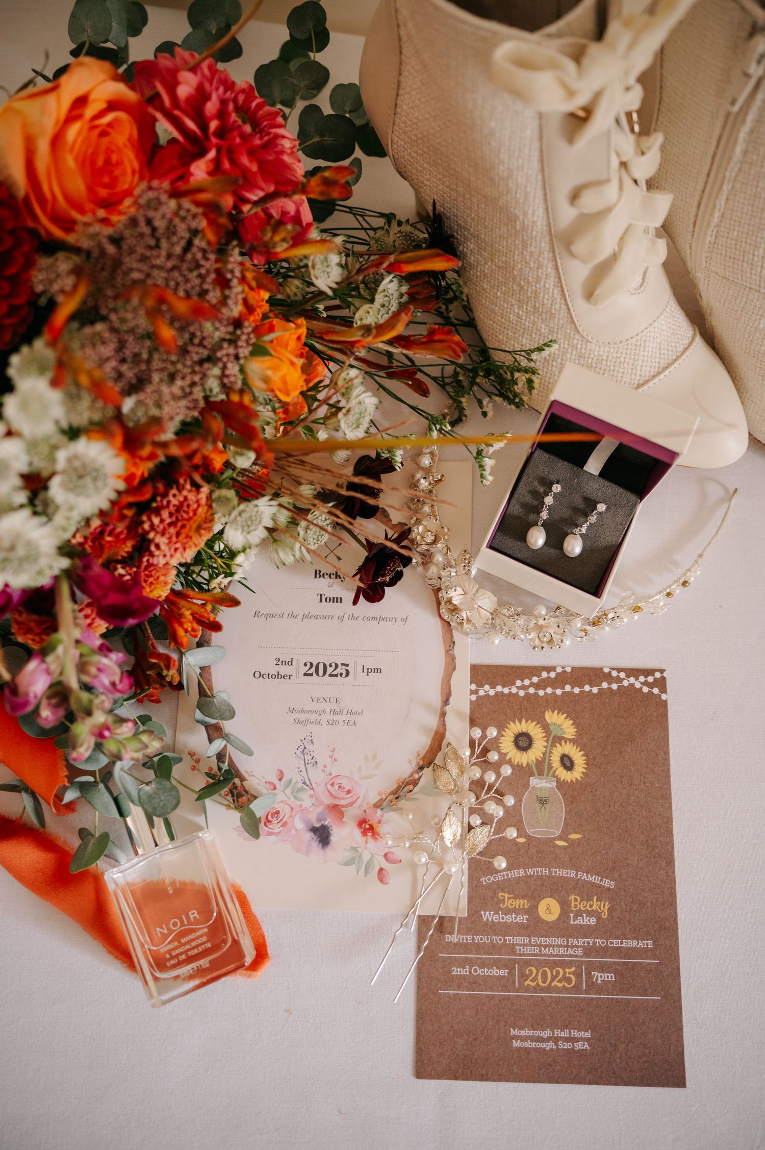 Wedding invitation suite with floral arrangements, pearls, and perfume bottle on a neutral background.