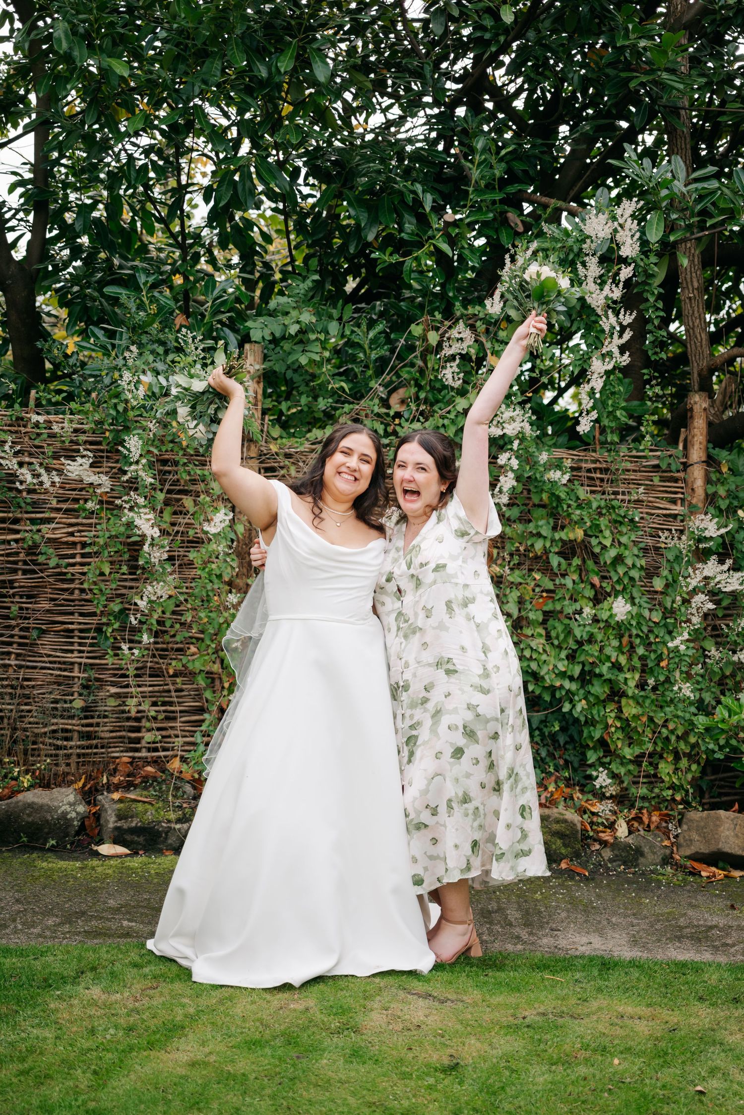 Two people in white dresses celebrating joyfully in a garden setting.