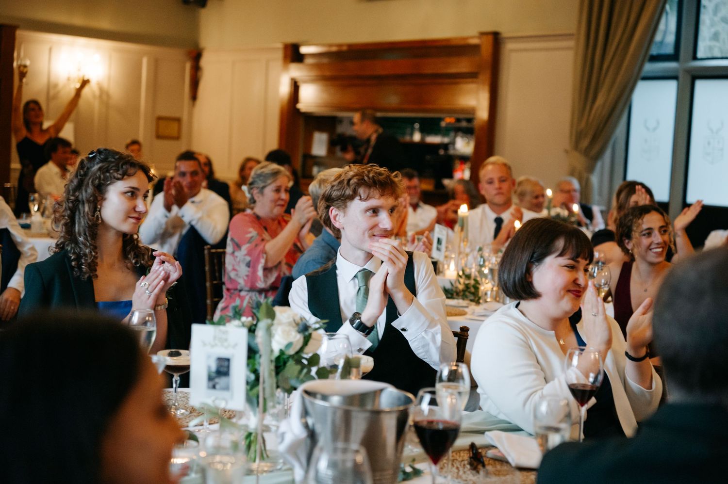 Wedding reception guests seated at long tables in an elegant indoor venue with warm lighting and decorative curtains.