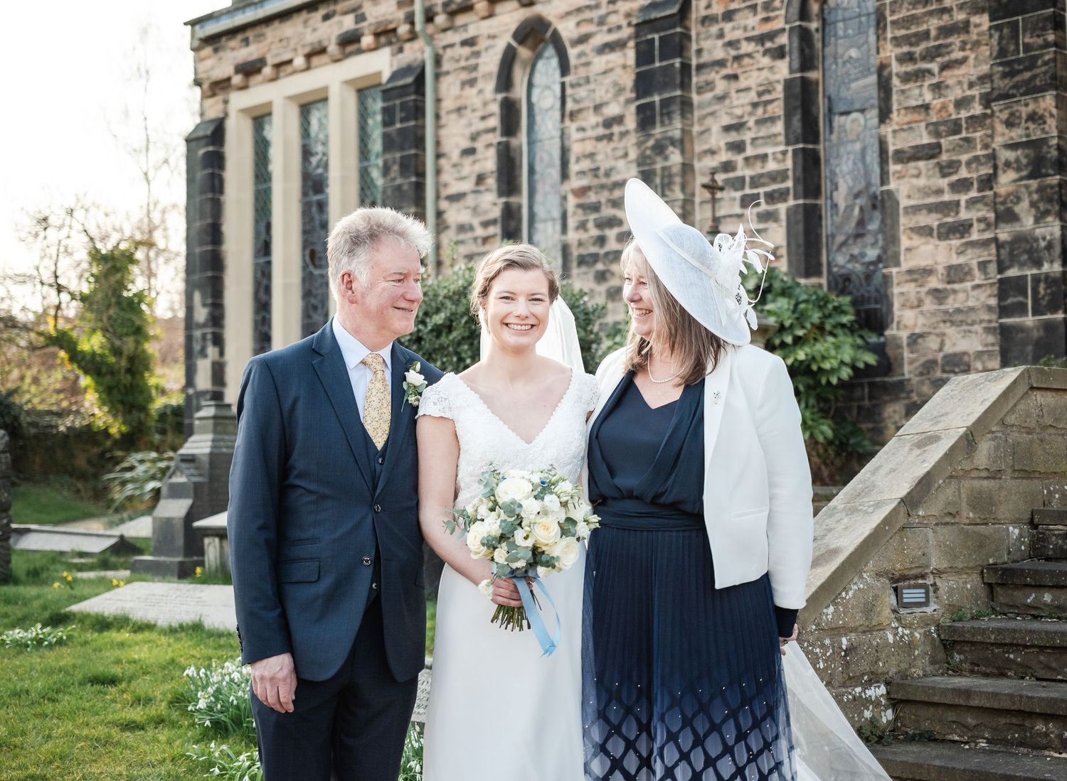 Wedding party poses outside a historic stone church building on a sunny day.
