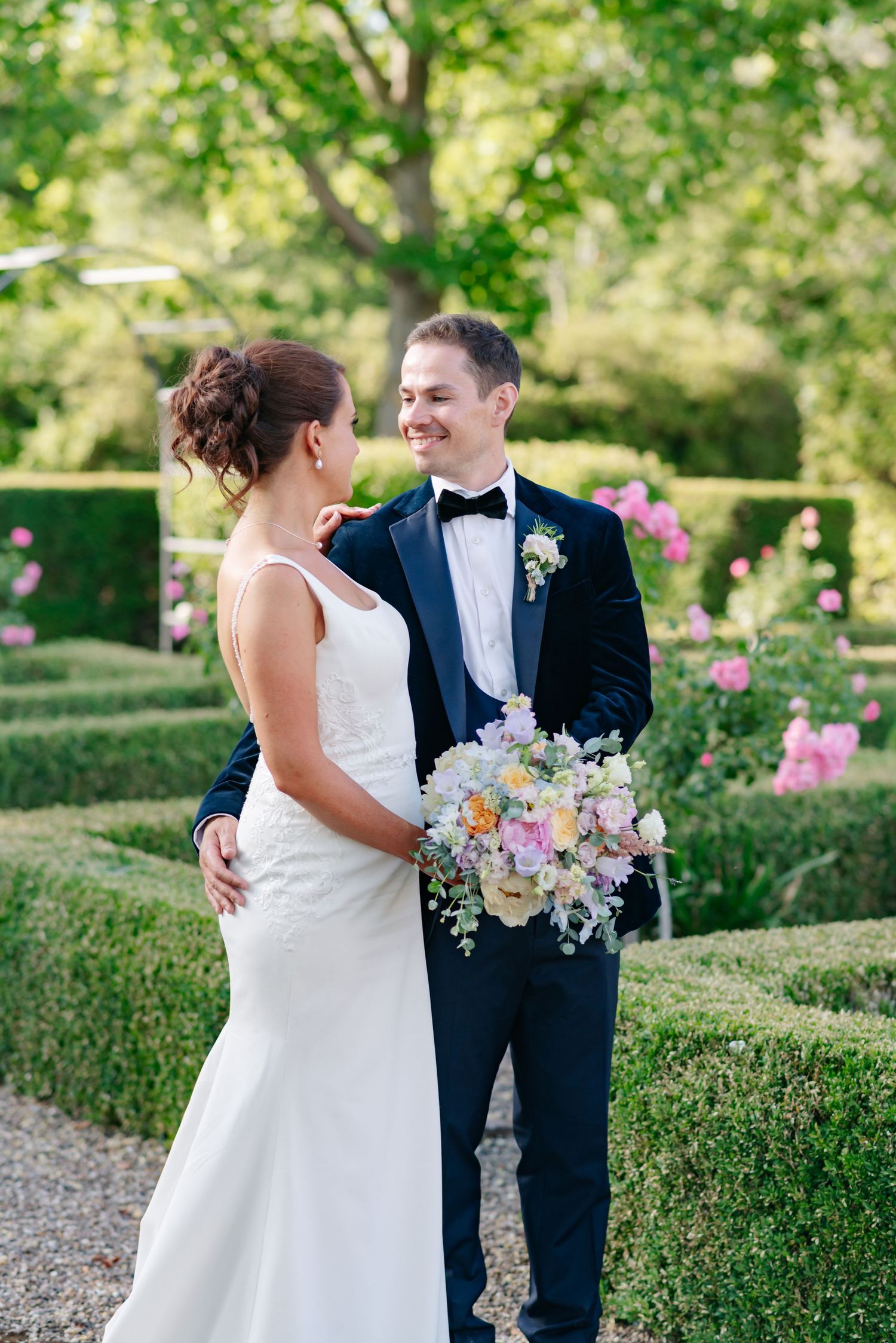 Wedding couple shares intimate moment in manicured garden with pink roses and hedges.