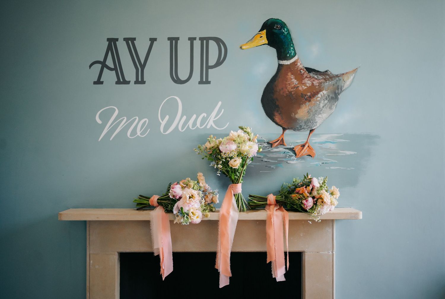 A duck decoy sits on a white shelf against a teal wall with floral arrangements.
