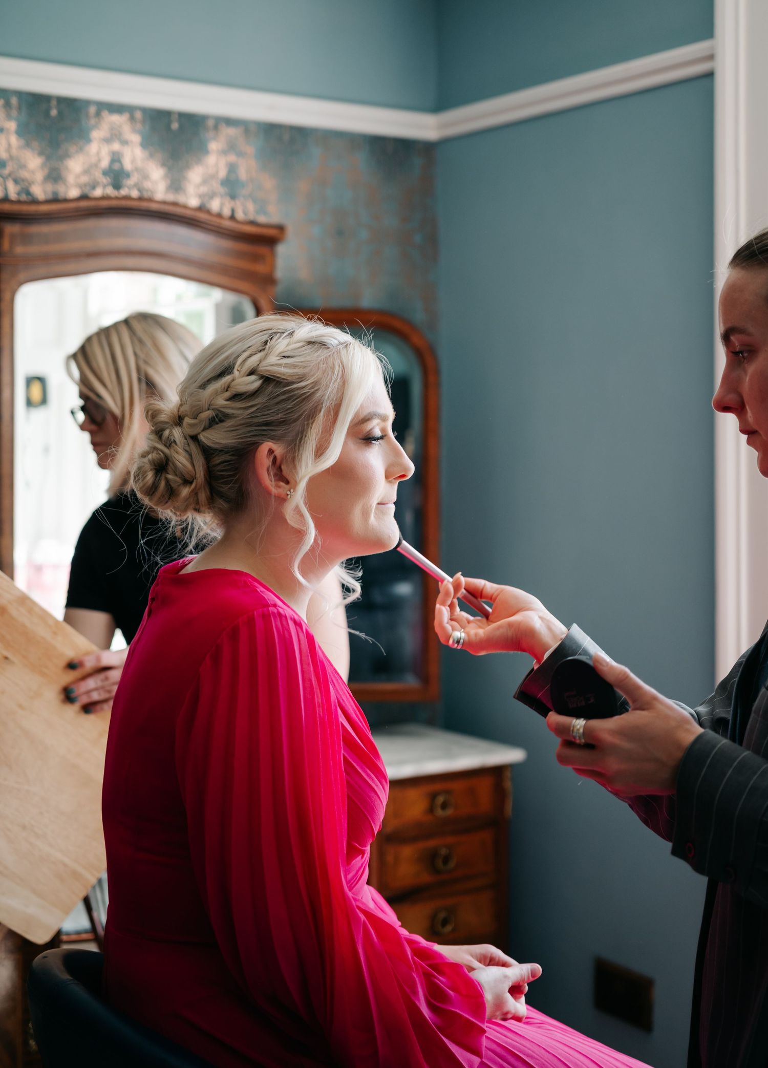 A wedding preparation scene shows someone getting makeup applied while wearing a vibrant red dress in an elegant room.