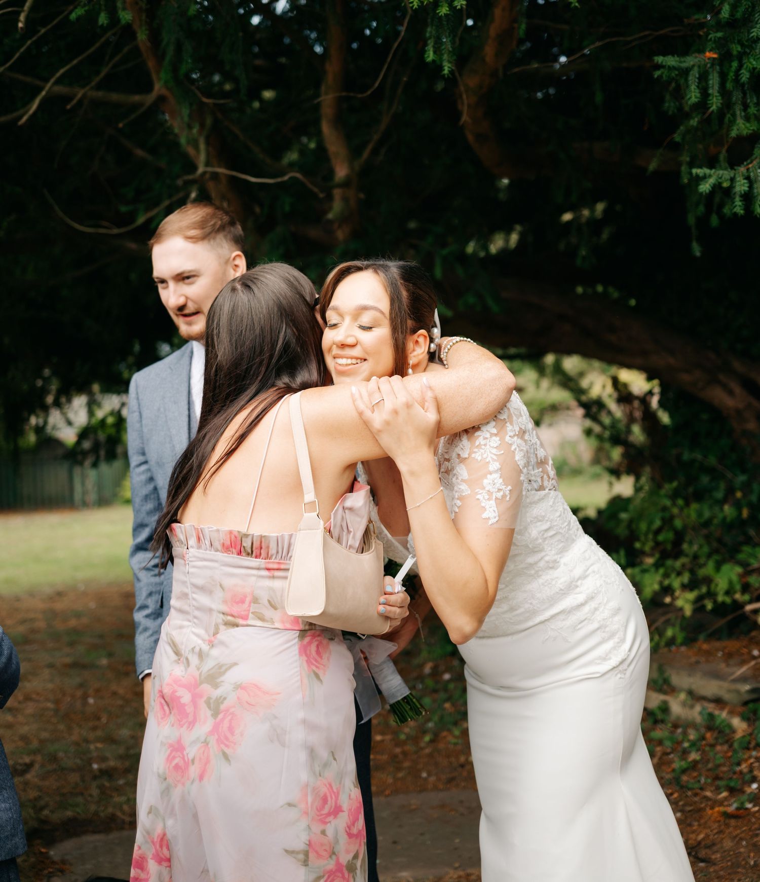 Emotional wedding moment with guests hugging in floral dresses outdoors.