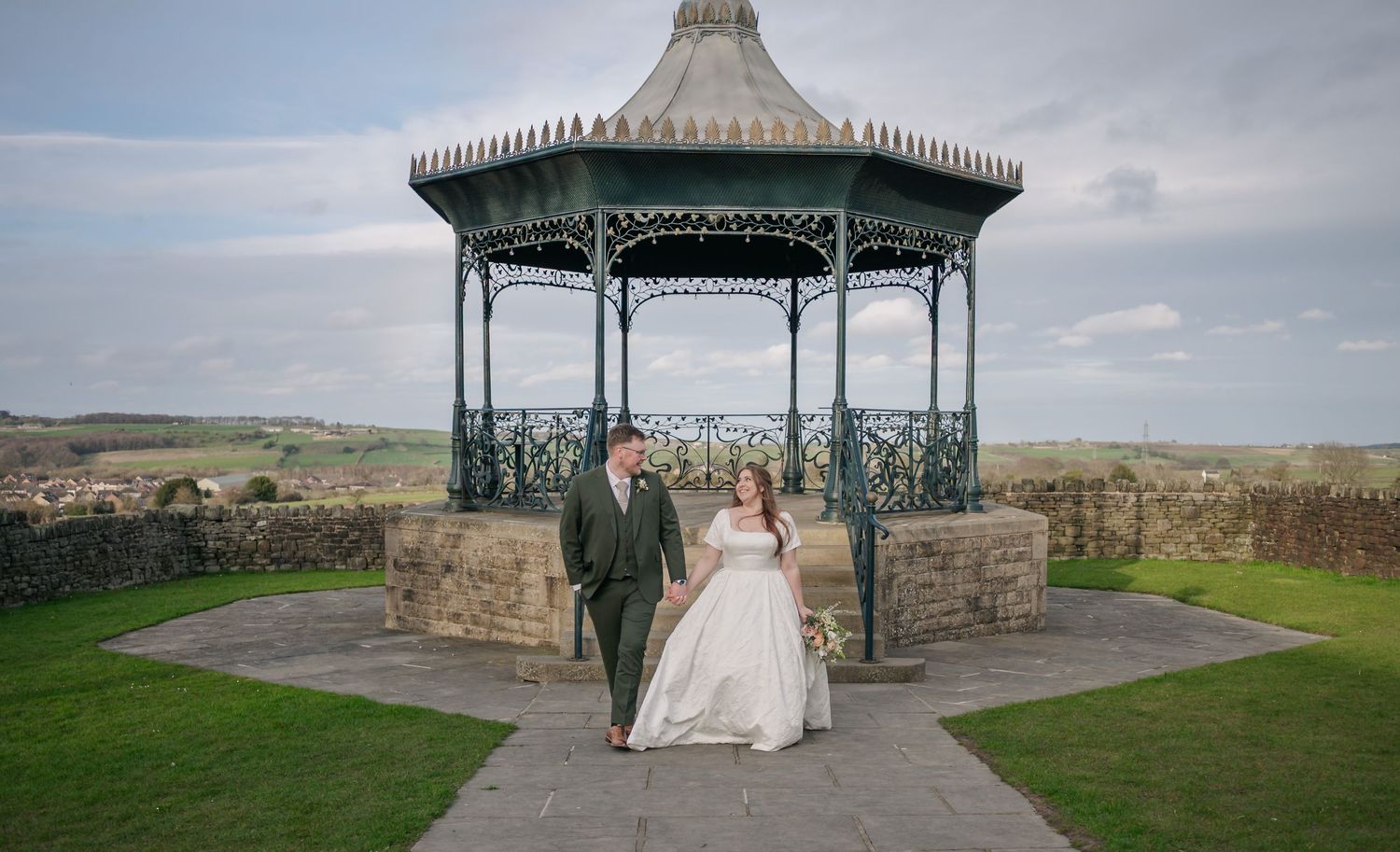 Couple standing in ornate Victorian-style bandstand gazebo overlooking scenic countryside landscape.