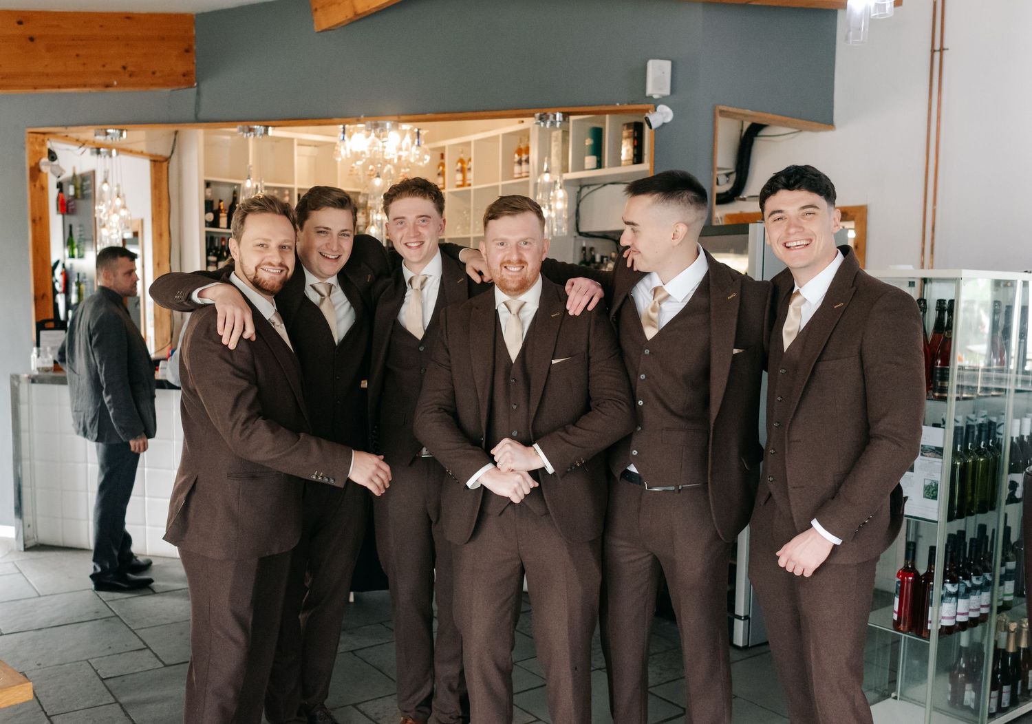 Groomsmen in matching brown suits pose together at a wedding venue with glass displays in the background.