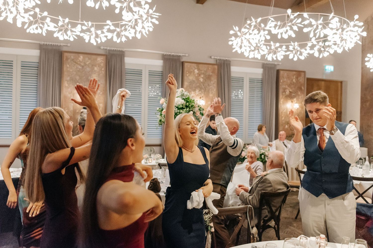 Wedding guests dancing and celebrating with raised arms under decorative white floral chandeliers.
