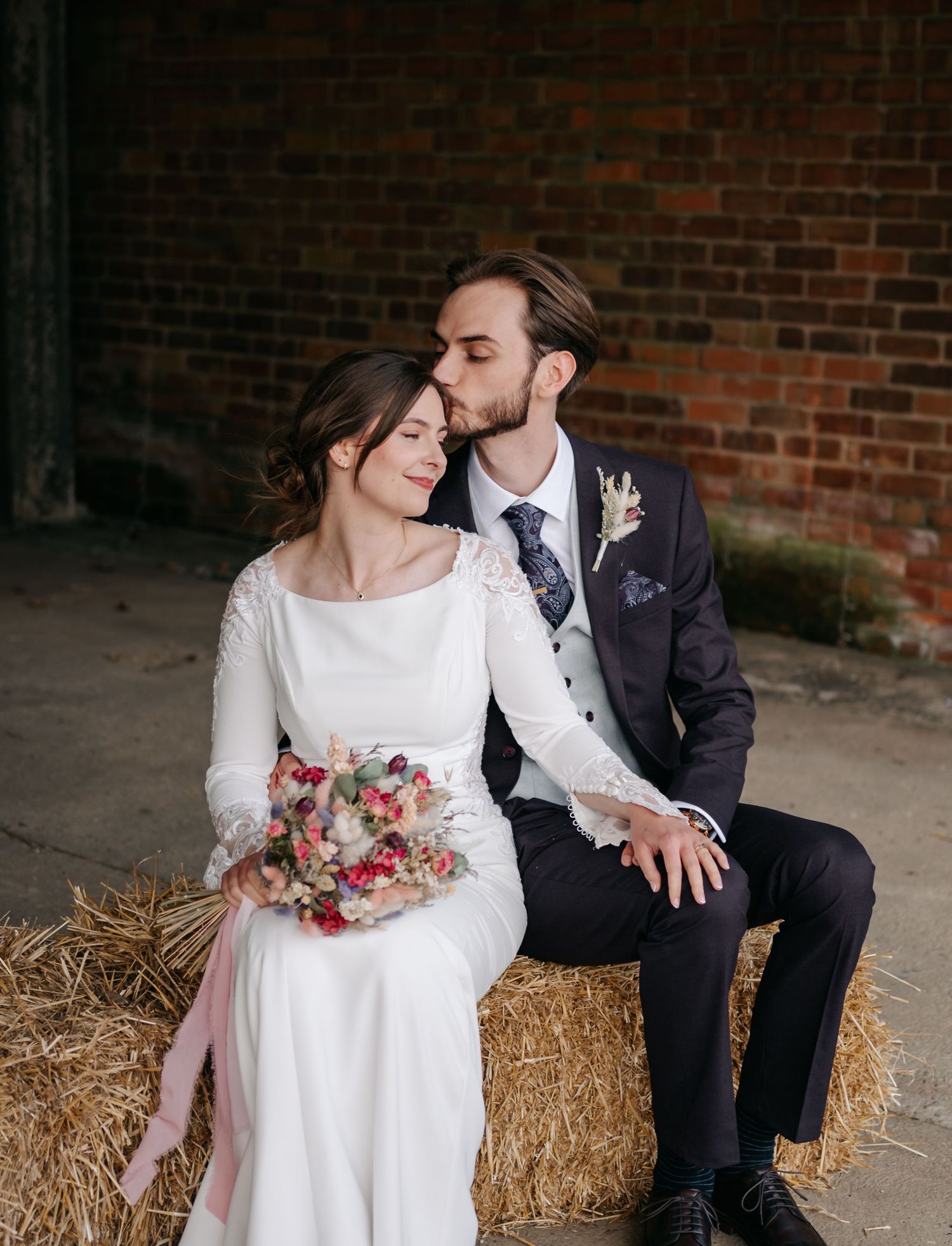 Newlyweds share intimate moments while seated on hay bales against brick wall backdrop.
