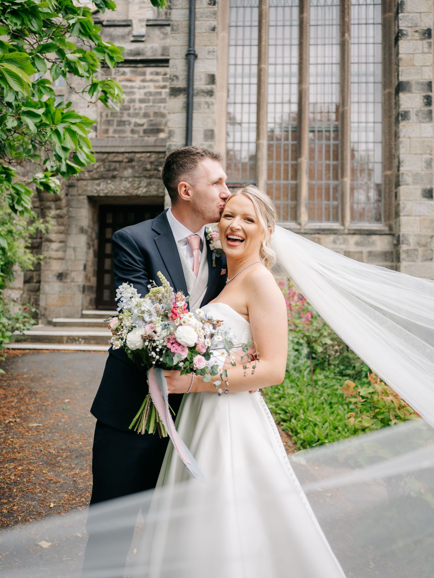 Newlyweds share a joyful moment outside a historic stone church with a flowing veil and pink floral bouquet.