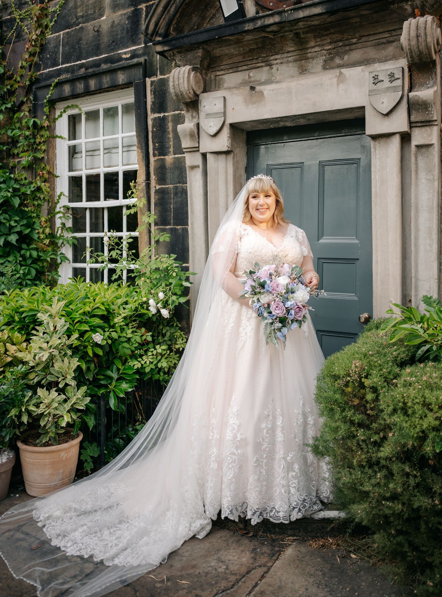Bride in lace ball gown with veil holds pastel bouquet outside stone building with teal door.