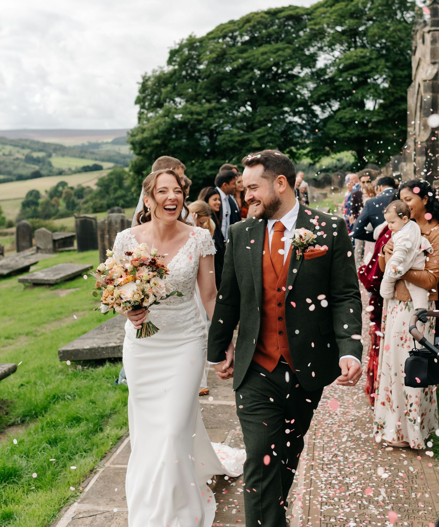 Smiling bride in a lace wedding gown and groom in a green suit walk through falling confetti outside a countryside church.