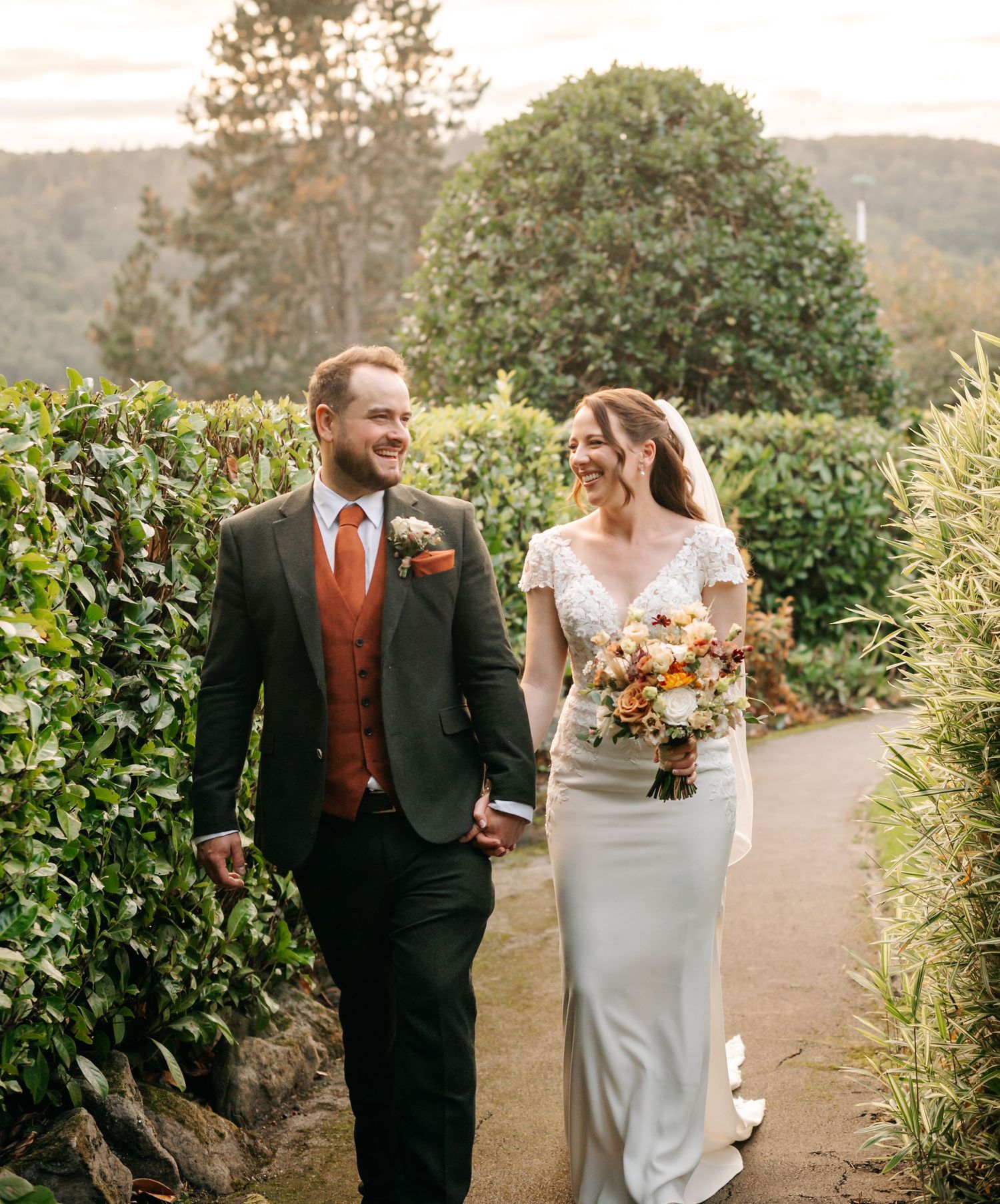Smiling bride and groom hold hands walking along a garden path lined with green hedges, she carries a colorful wedding bouquet.
