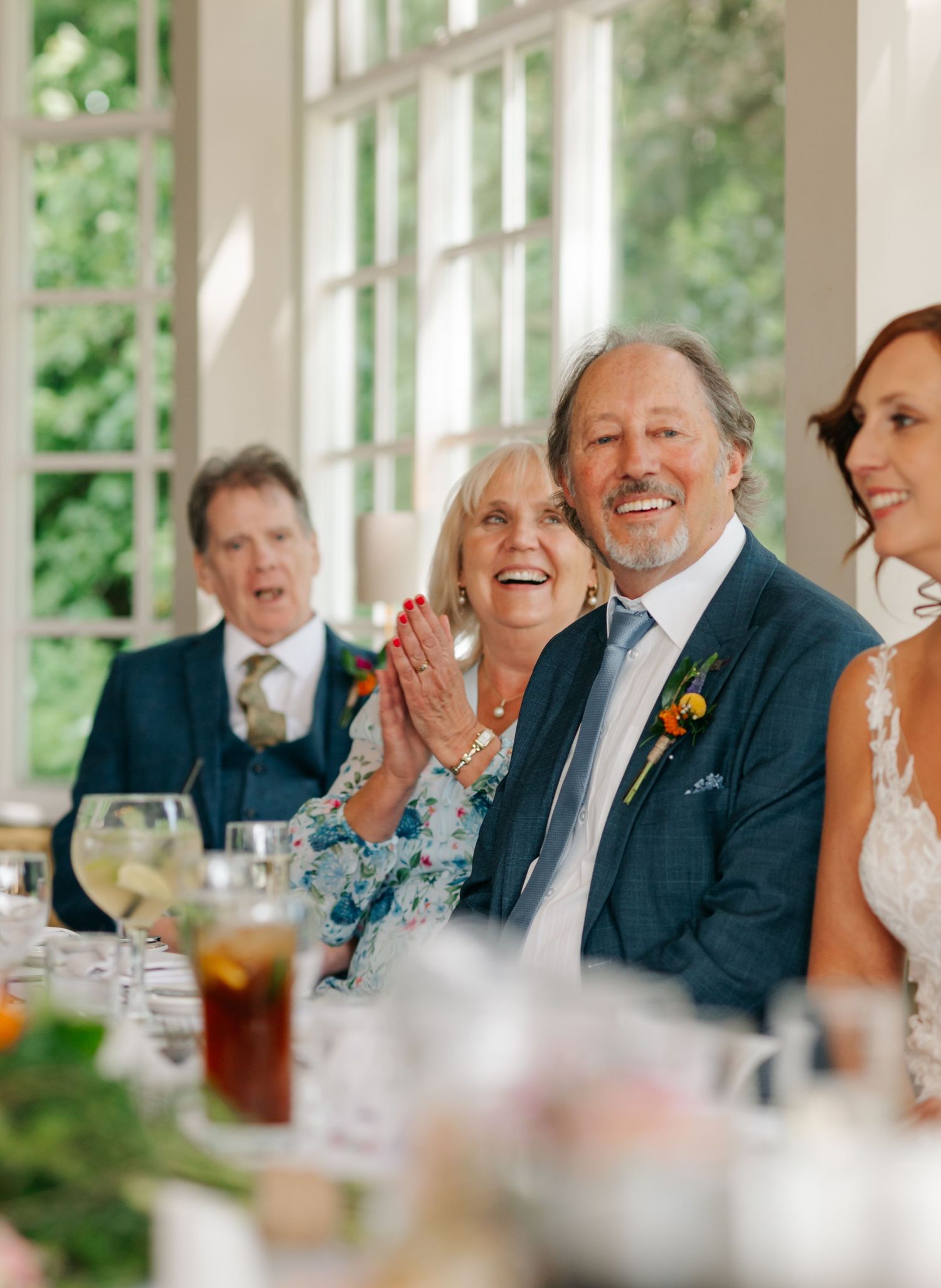 Wedding guests share laughter and joy during reception dinner at bright sunlit venue with green foliage visible outside.