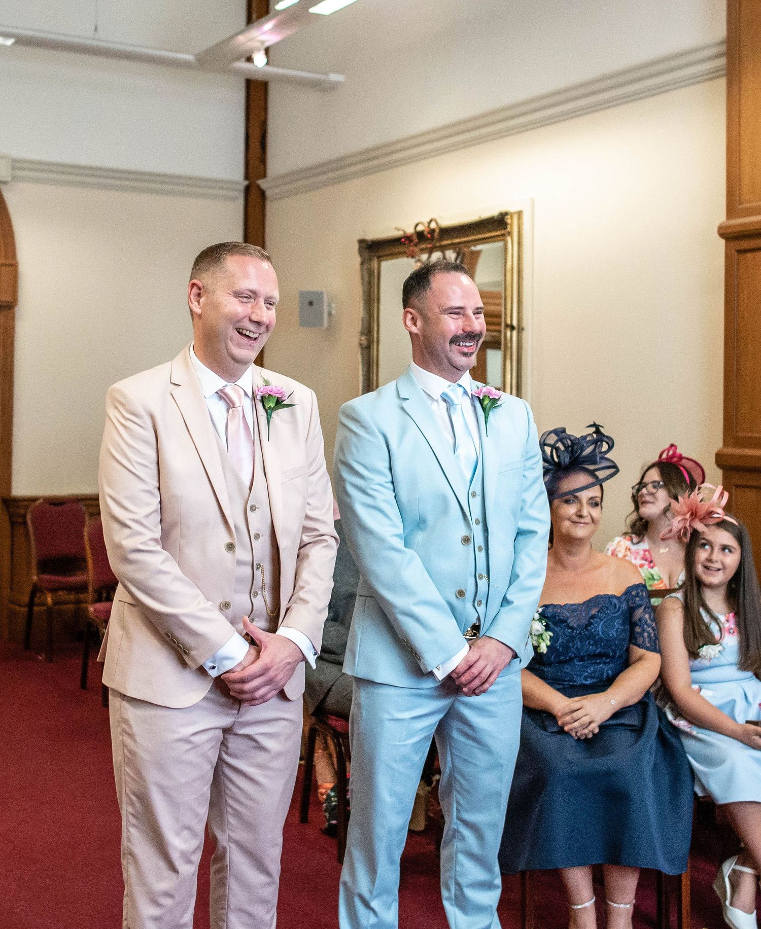 Wedding party stands in a reception room with guests in pink and blue suits at the front.