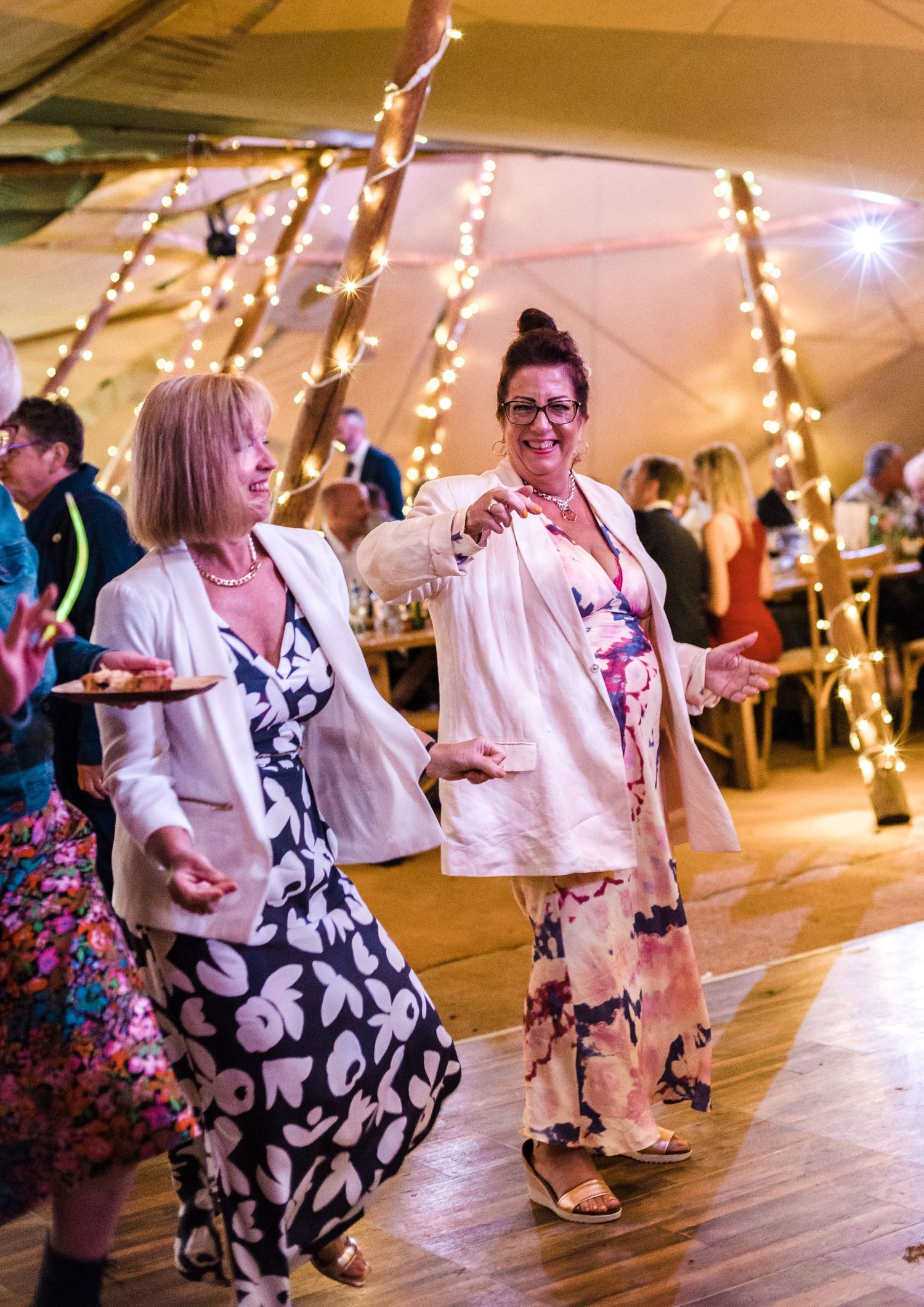 Two guests dancing at a wedding reception under string lights with elegant dresses and joyful expressions.