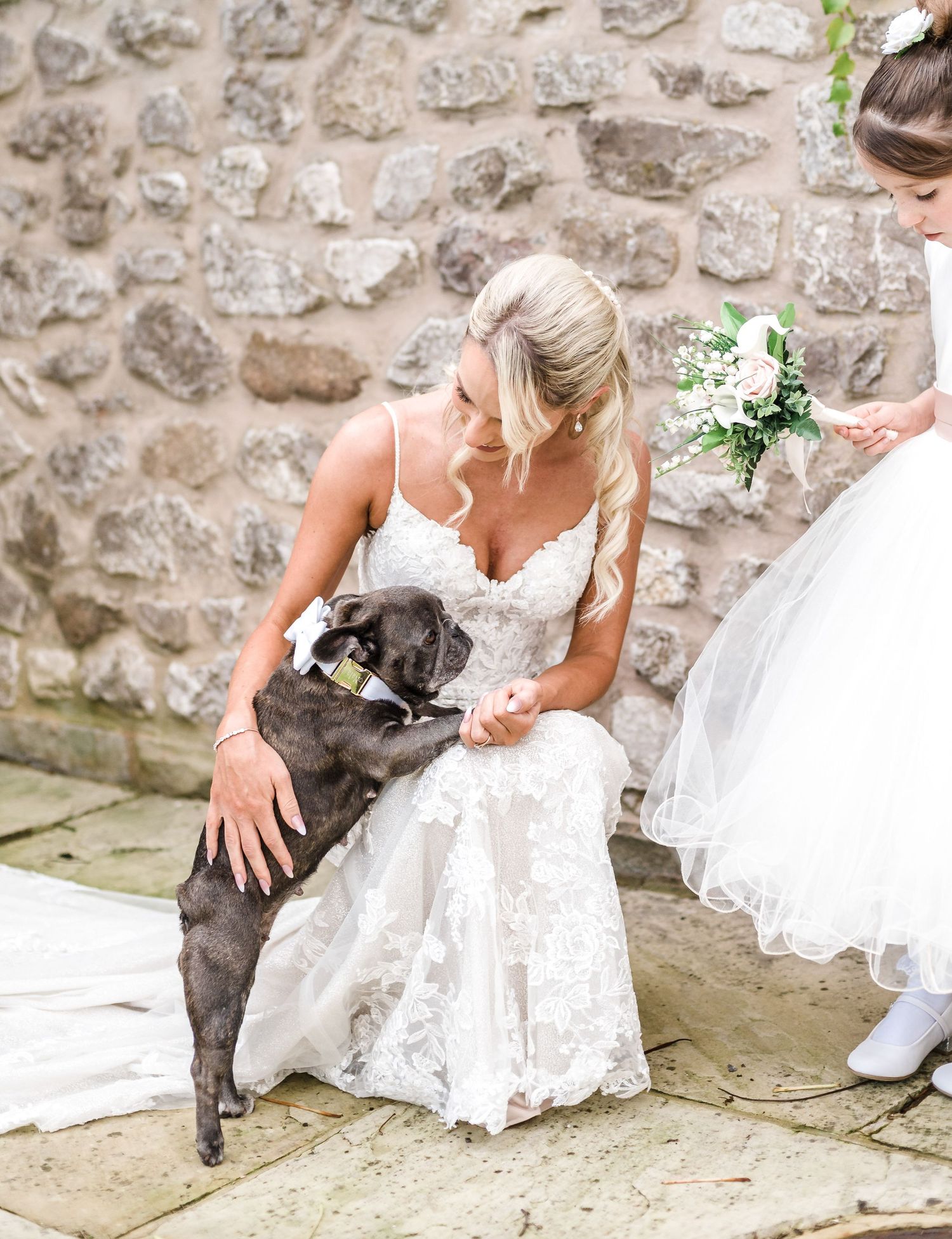 Touching wedding moment with a gray dog and flowing white dress against stone wall.