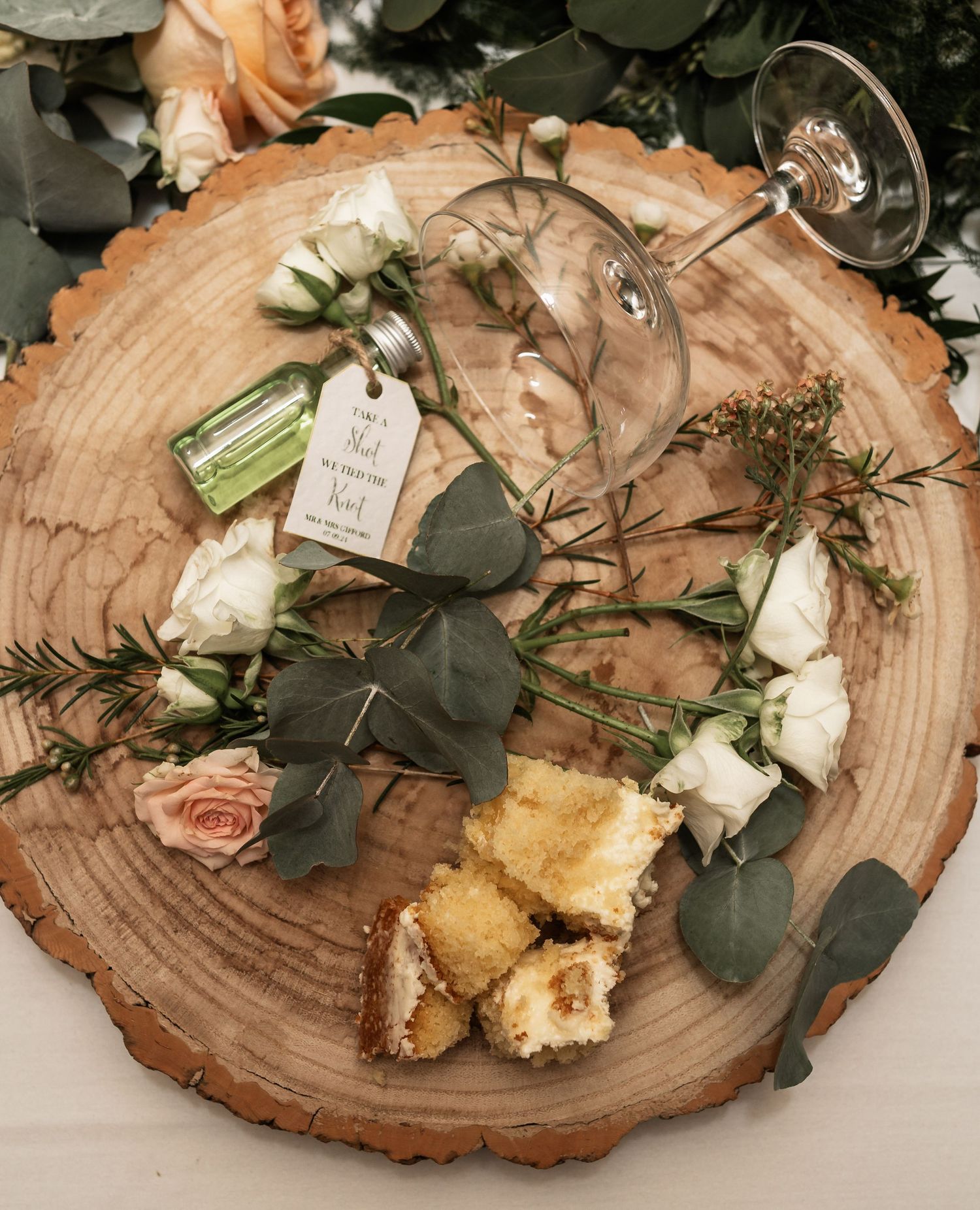 Wedding details arranged on a rustic wood slice including wine glasses, eucalyptus, and small cake pieces.