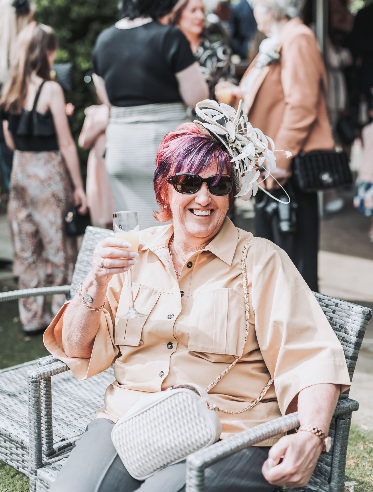 Wedding guest in beige outfit and fascinator smiles while holding drink at outdoor reception.