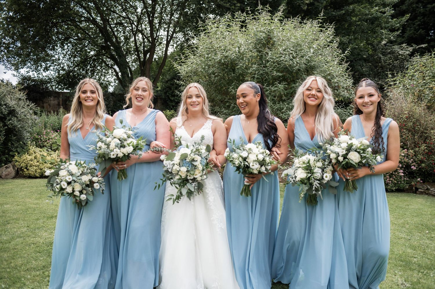 Wedding party in matching powder blue dresses poses with bouquets in garden setting.