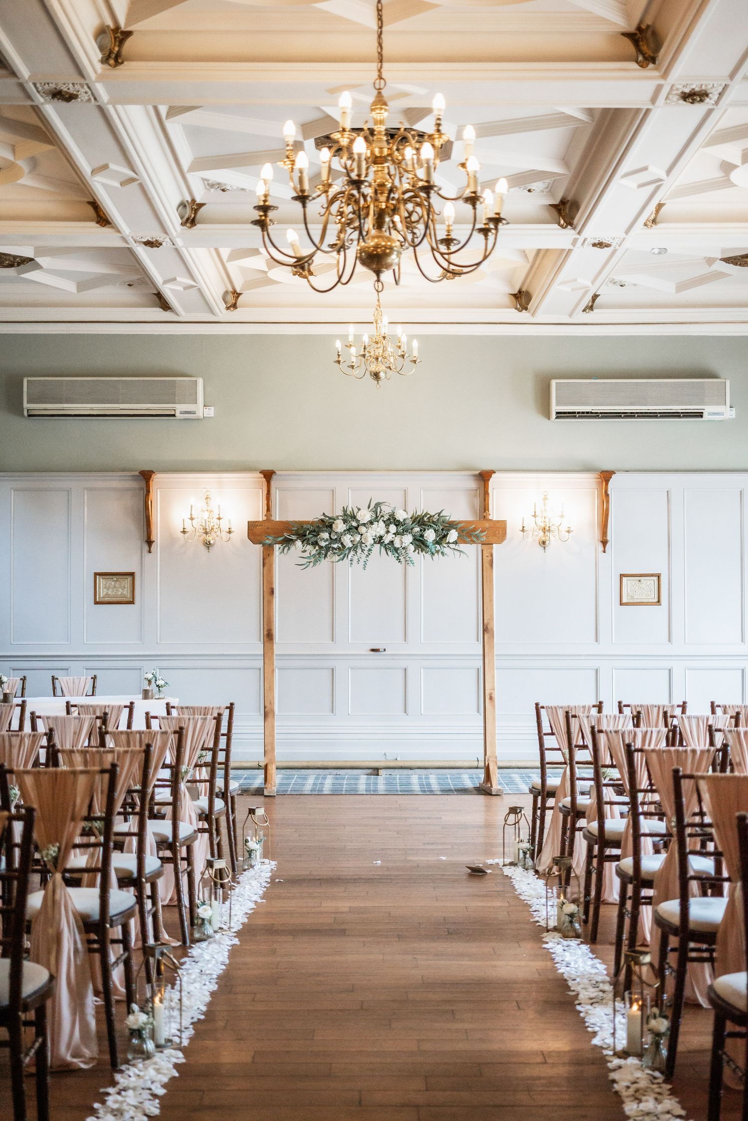 Wedding ceremony setup with wooden chairs and floral arch under elegant chandeliers.