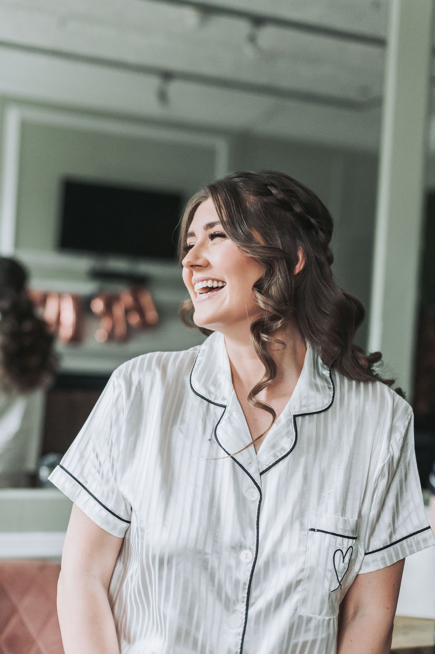 A person wearing white pajamas with black piping laughs joyfully during wedding preparations.