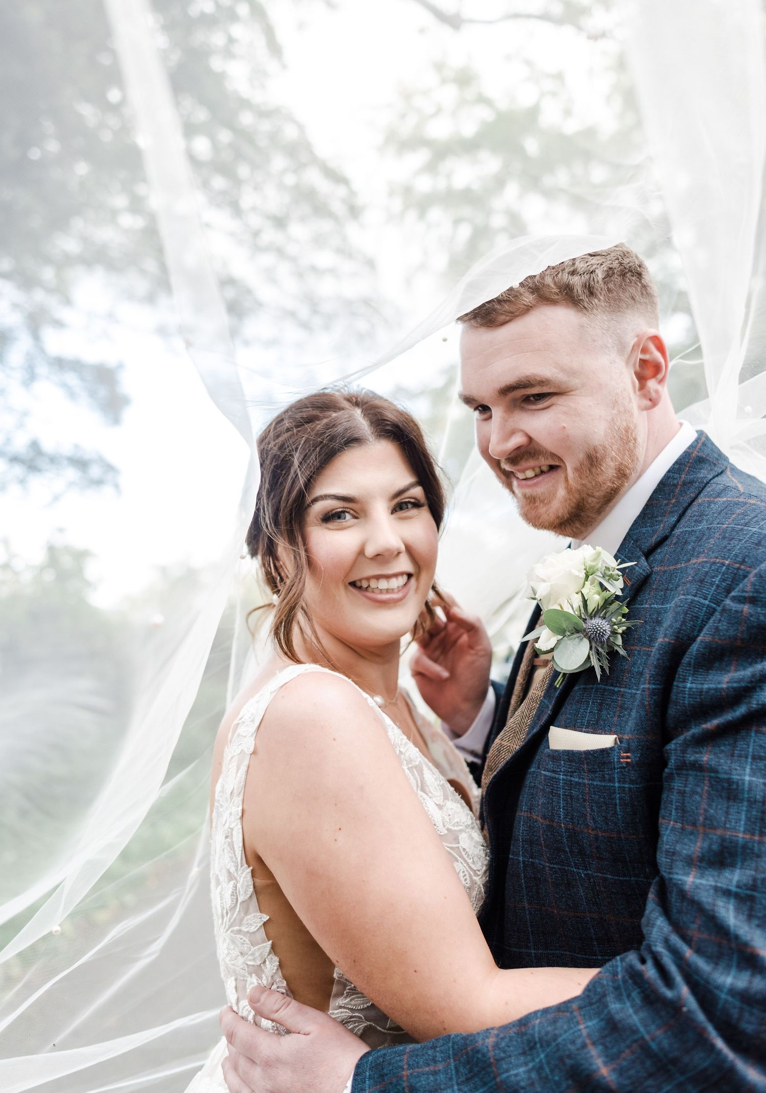 Joyful wedding portrait with bride's veil floating in the breeze against natural background.