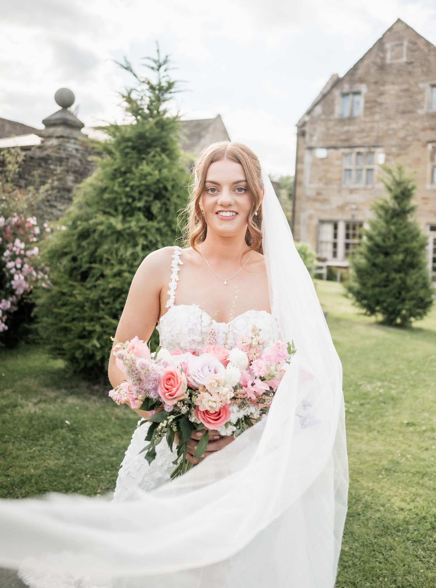 Bride in white gown holds pink and purple bouquet in front of stone mansion with flowing veil.