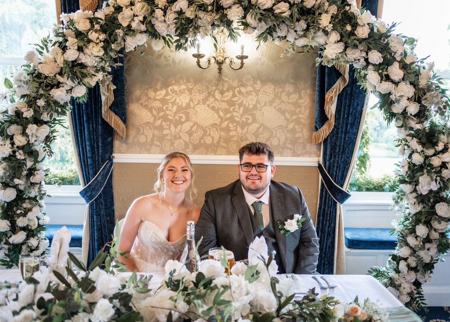 Newlyweds seated under beautiful floral arch with white and pink roses.