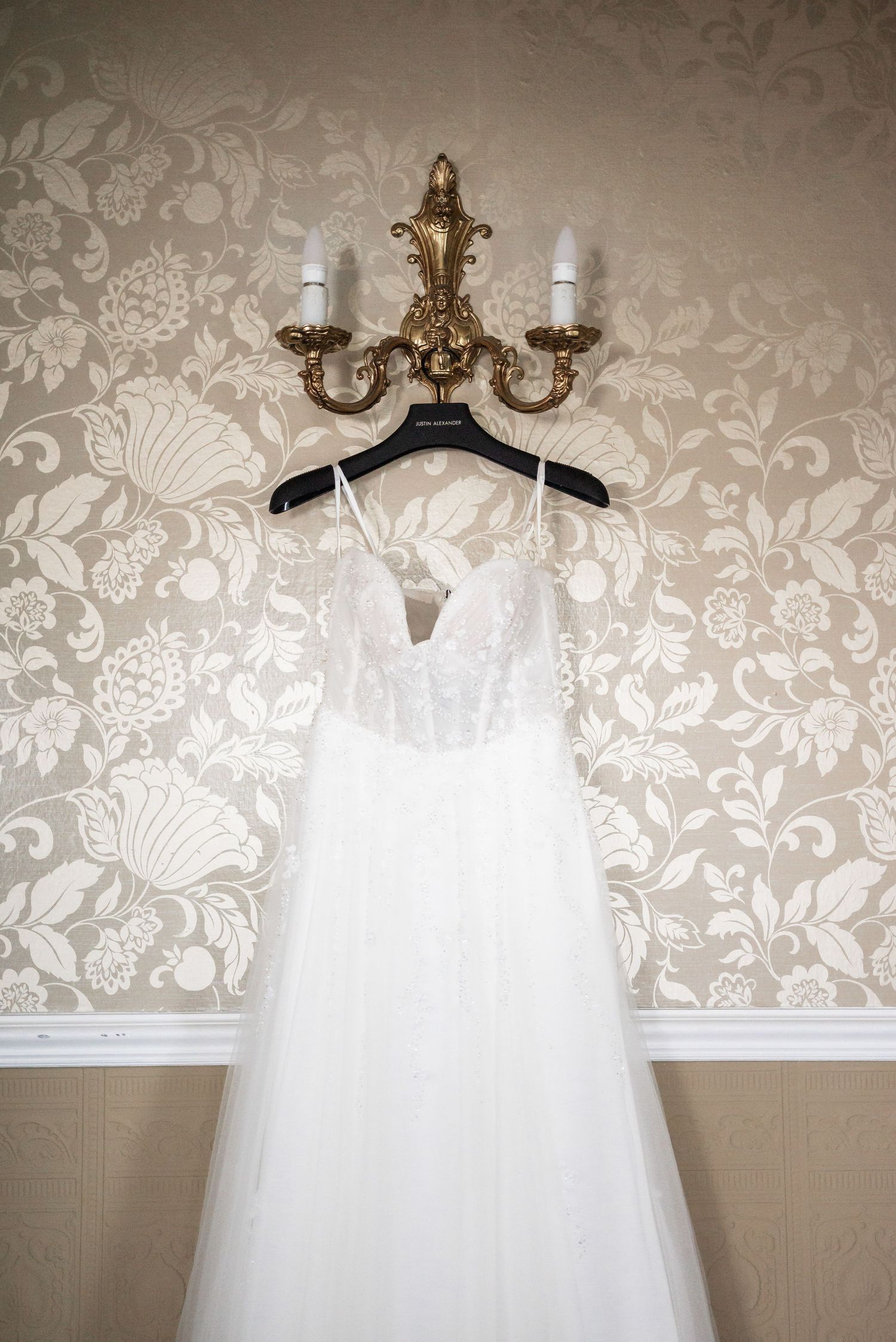 White wedding dress hanging on ornate gold chandelier against floral wallpaper.