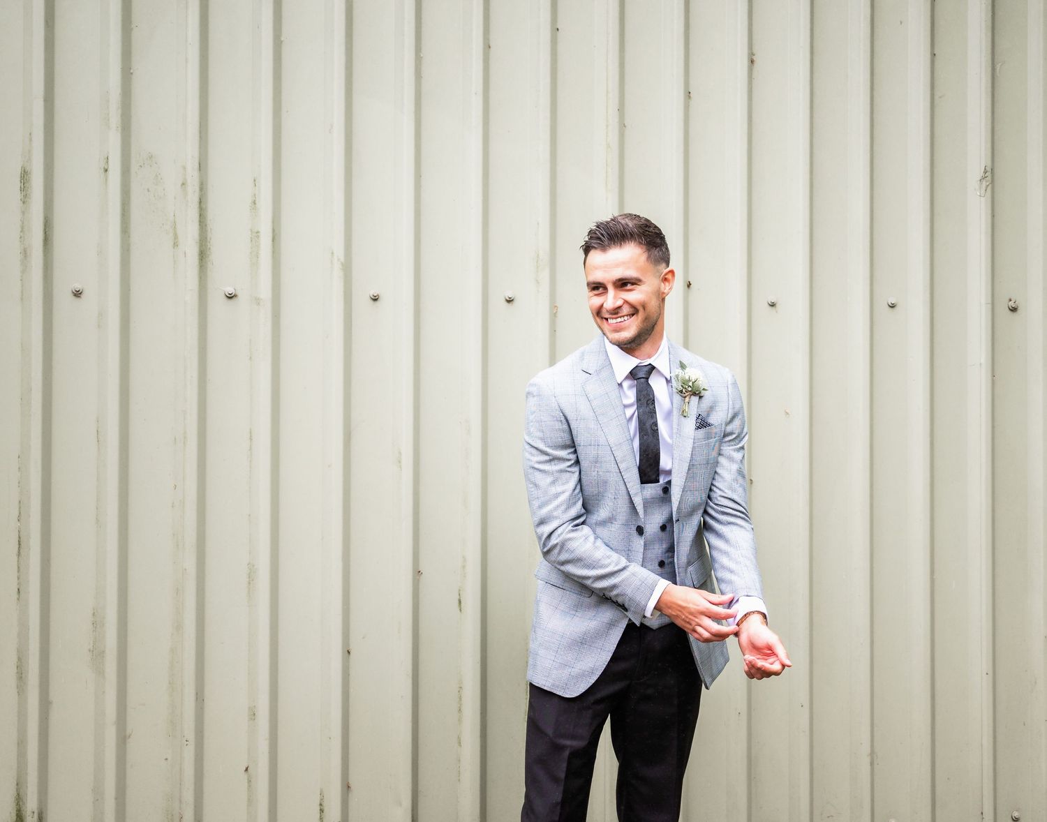 Man in light gray suit jacket and tie poses against corrugated metal background.