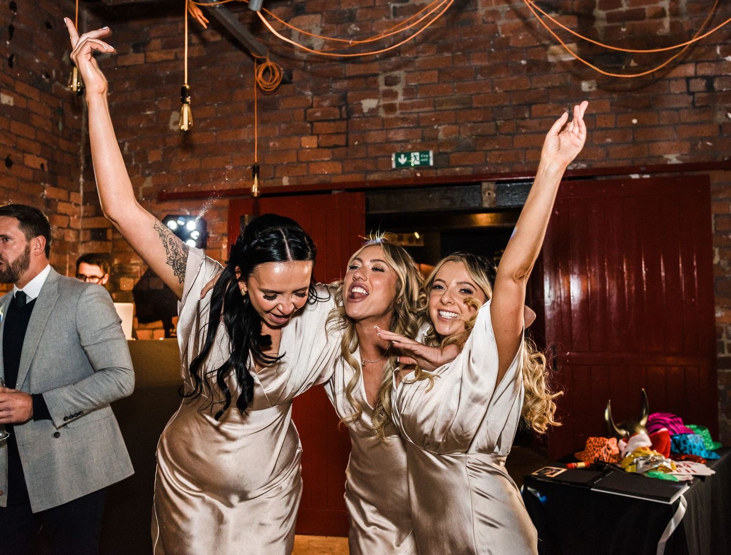 Bridesmaids in silk dresses dancing and celebrating with arms raised at wedding reception.