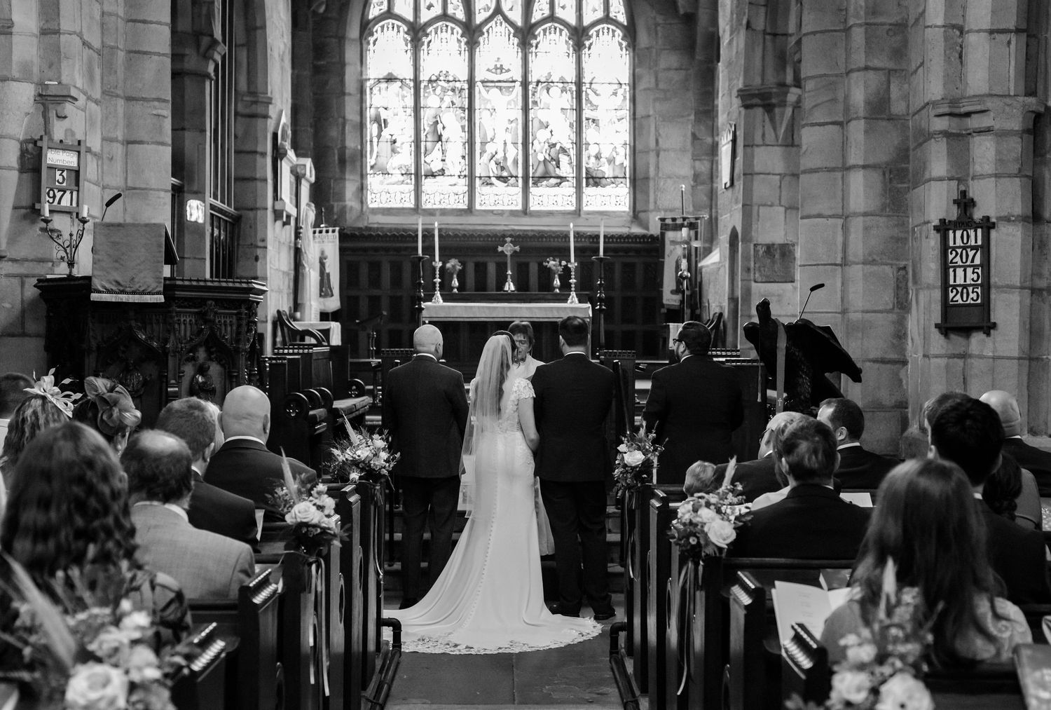 A black and white photo of a bride and groom standing at the altar of a church as wedding guests watch from the pews.