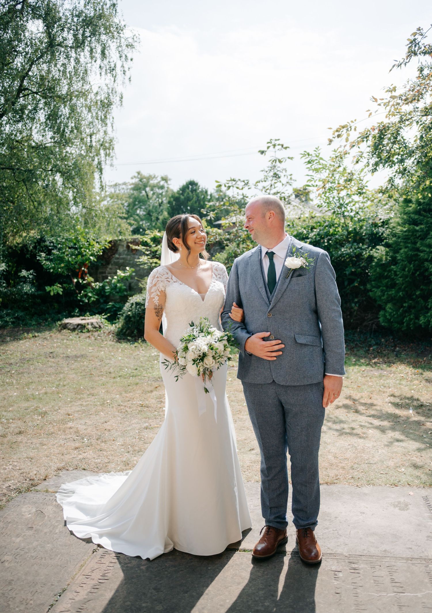 A couple in wedding attire poses outdoors with greenery in the background.