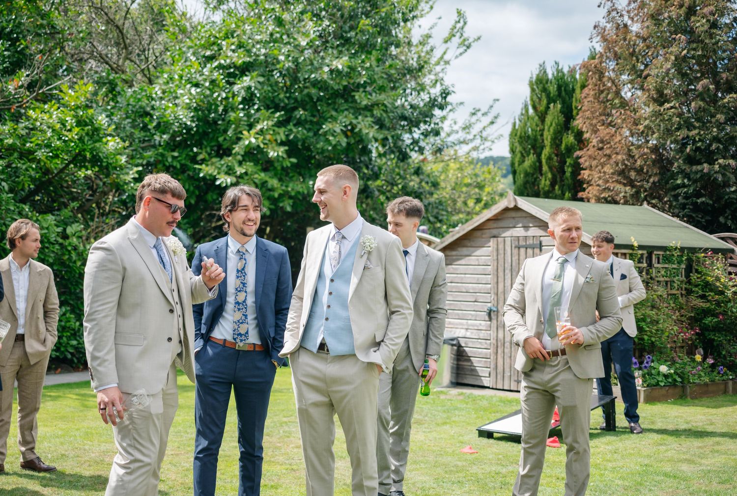 A group of groomsmen in light colored suits chat casually on a sunny lawn with a garden shed in the background.