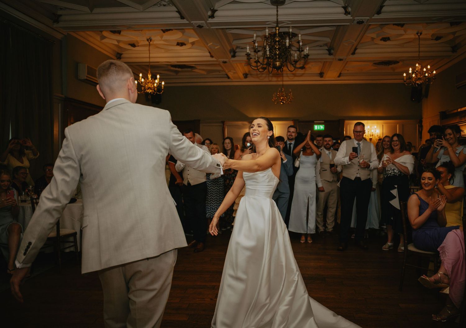 Wedding couple dancing on wooden floor surrounded by guests at dimly lit reception.