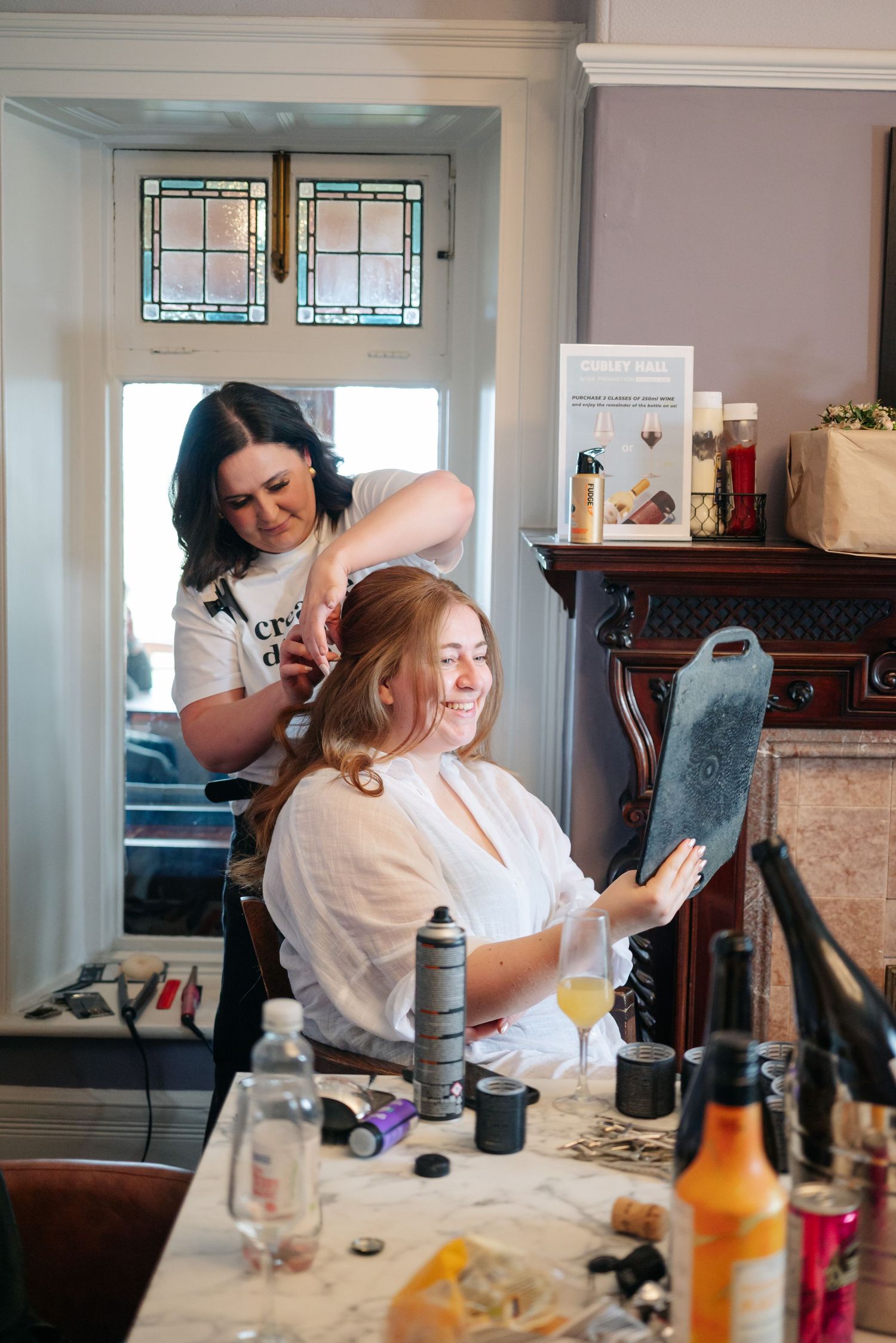 A wedding preparation scene showing hair styling while the client checks her phone in a room with stained glass windows.