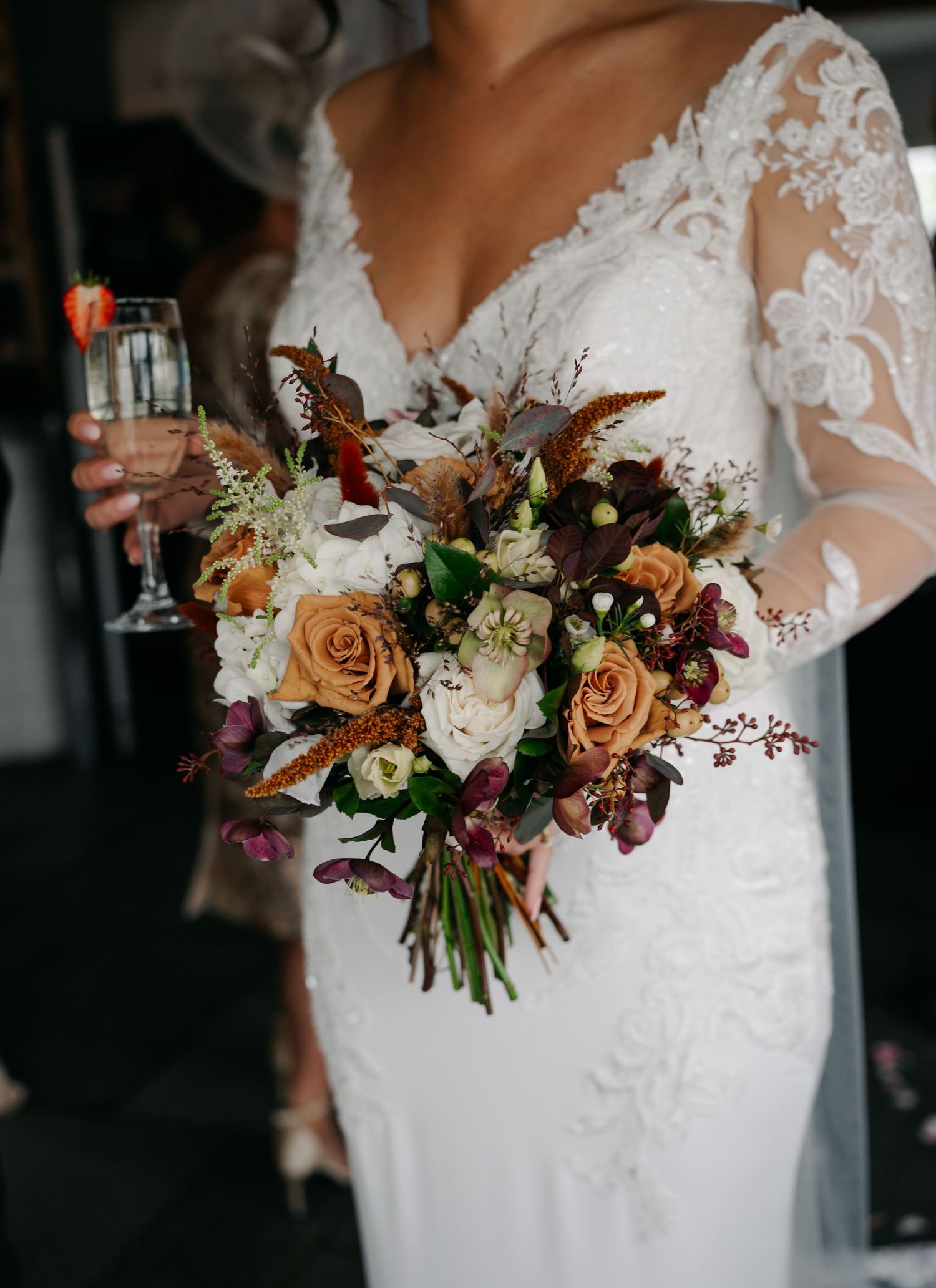 Close-up of a bridal bouquet with peach roses, white flowers and burgundy accents against a white lace wedding dress.